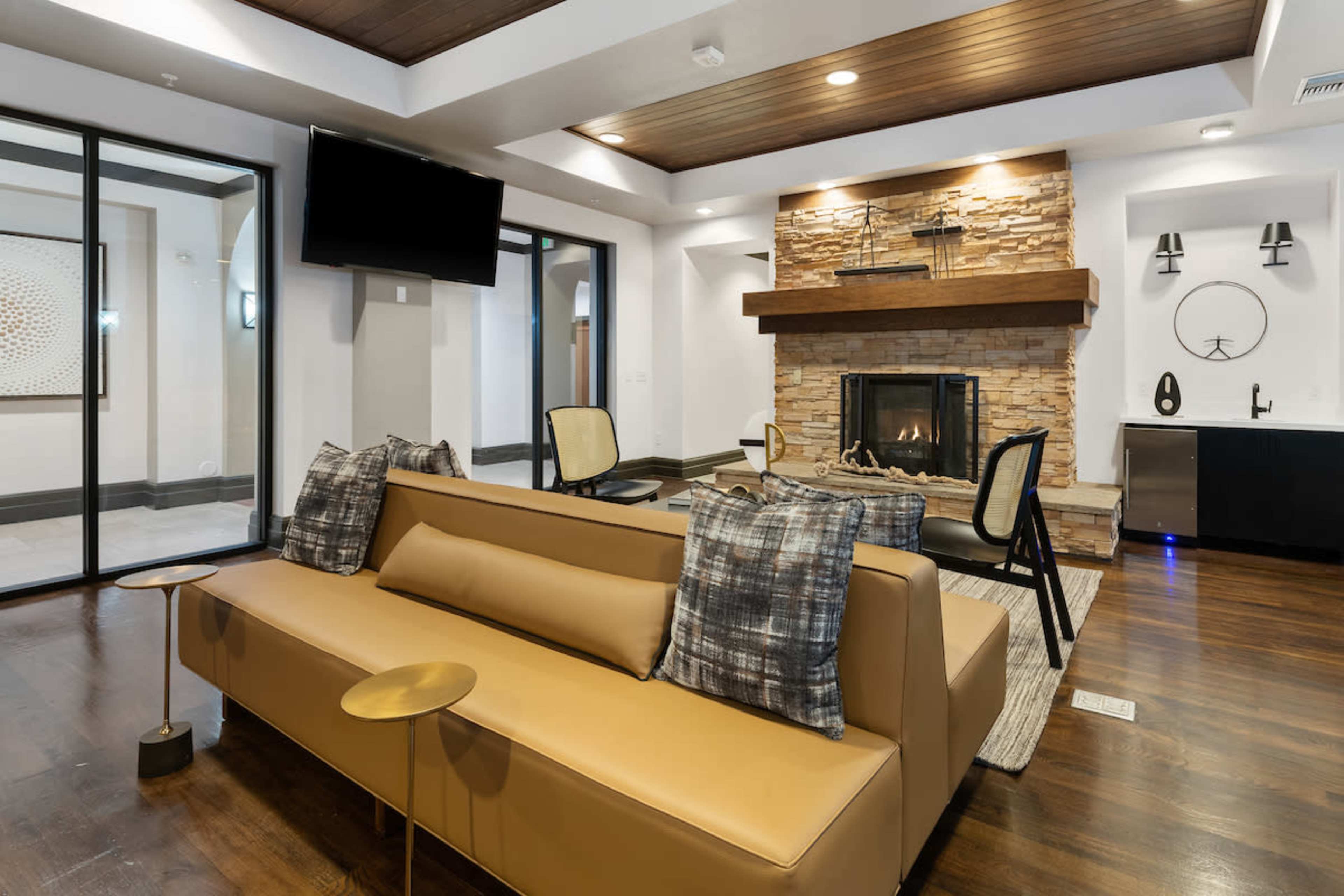 A modern living area featuring a tan sofa with decorative pillows, a stone fireplace, a wall-mounted TV, and large windows.