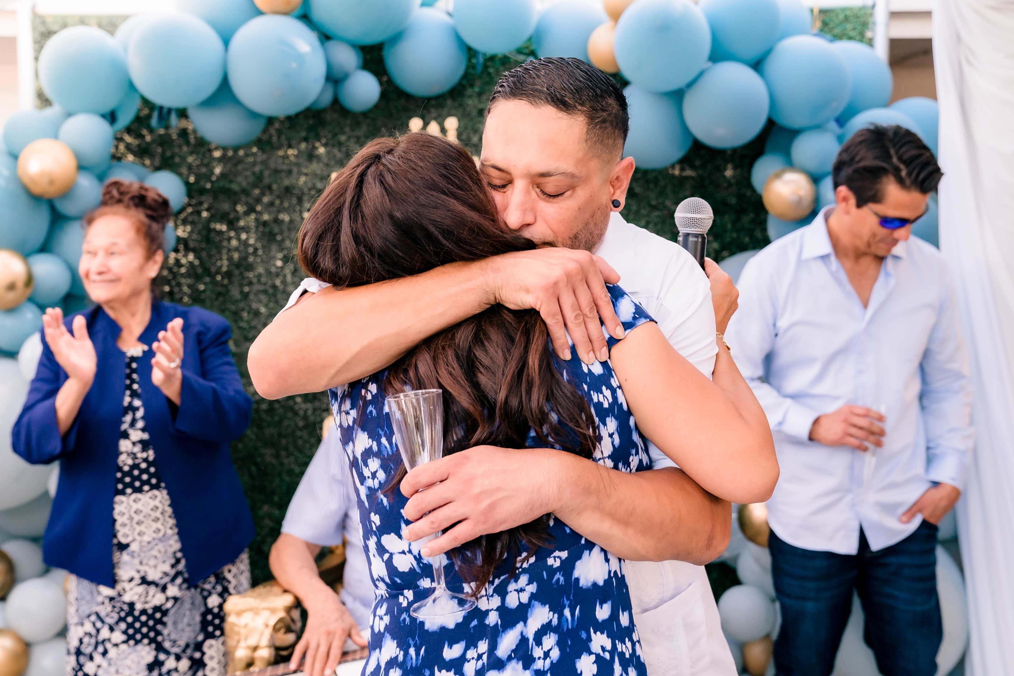 A man and woman embrace in celebration, surrounded by guests who are clapping and enjoying the event, with a backdrop of blue and gold balloons.