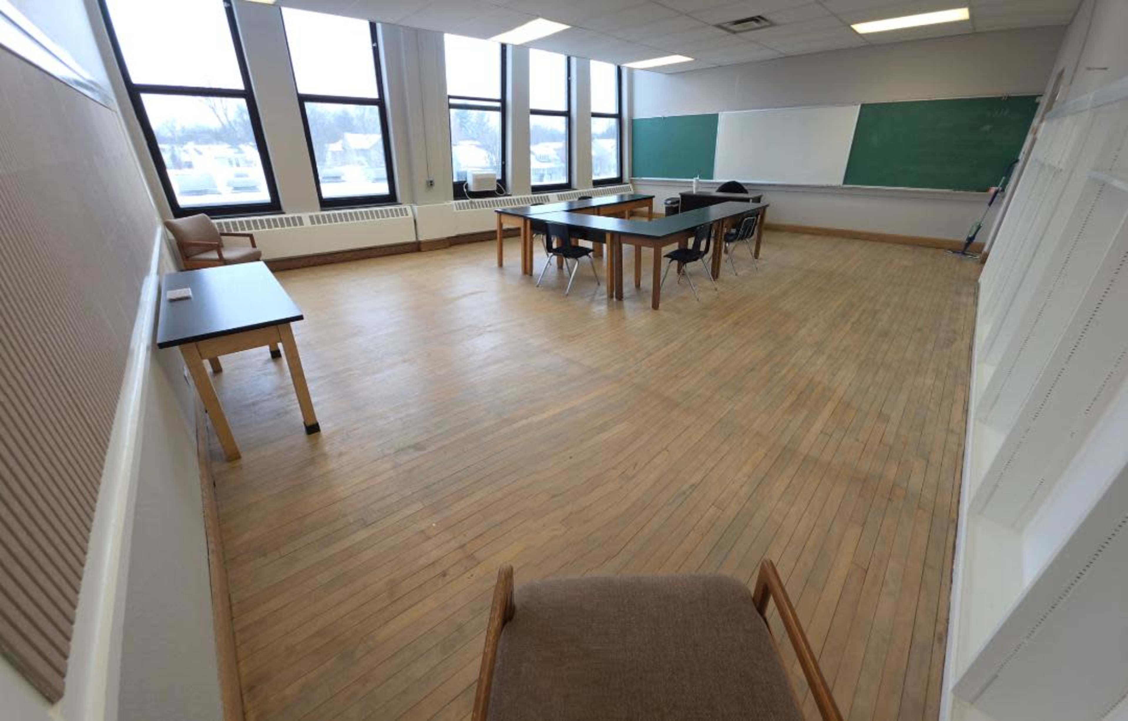 The image shows a classroom with multiple tables, chairs, and a chalkboard, featuring large windows that allow for natural light.