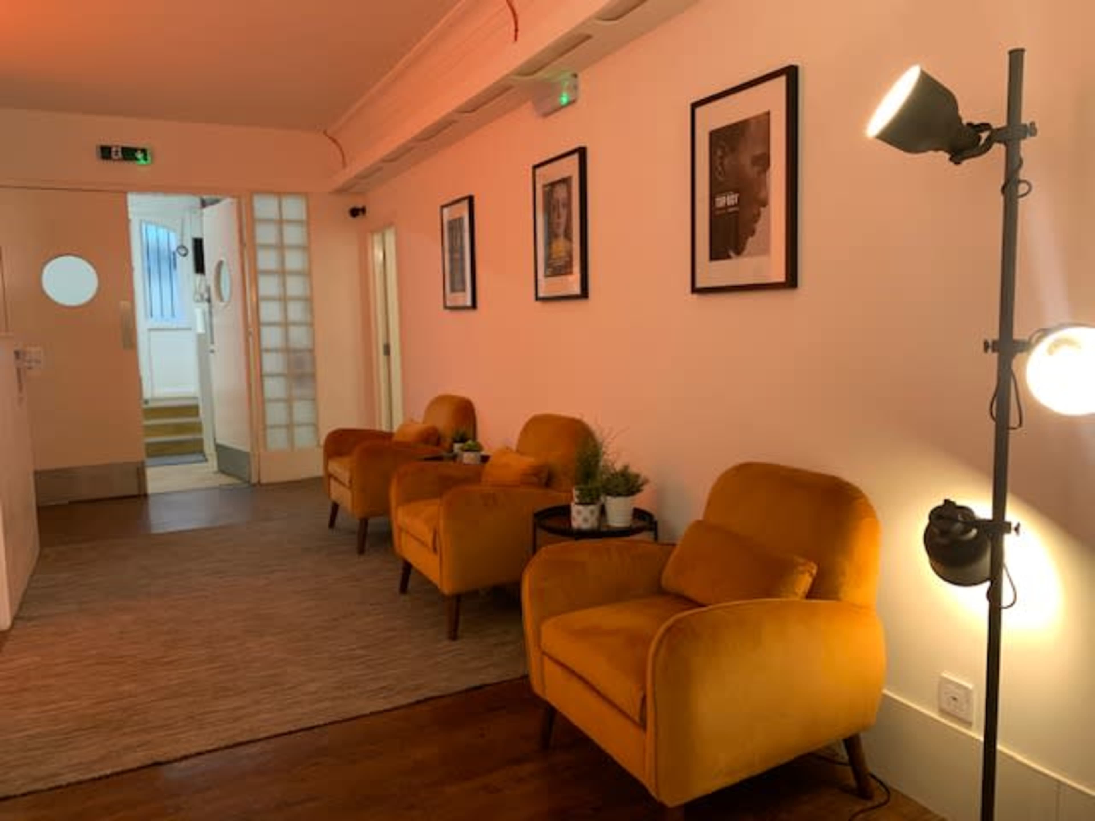 A hallway with three yellow chairs, small tables with plants, framed pictures on the wall, and soft lighting fixtures.