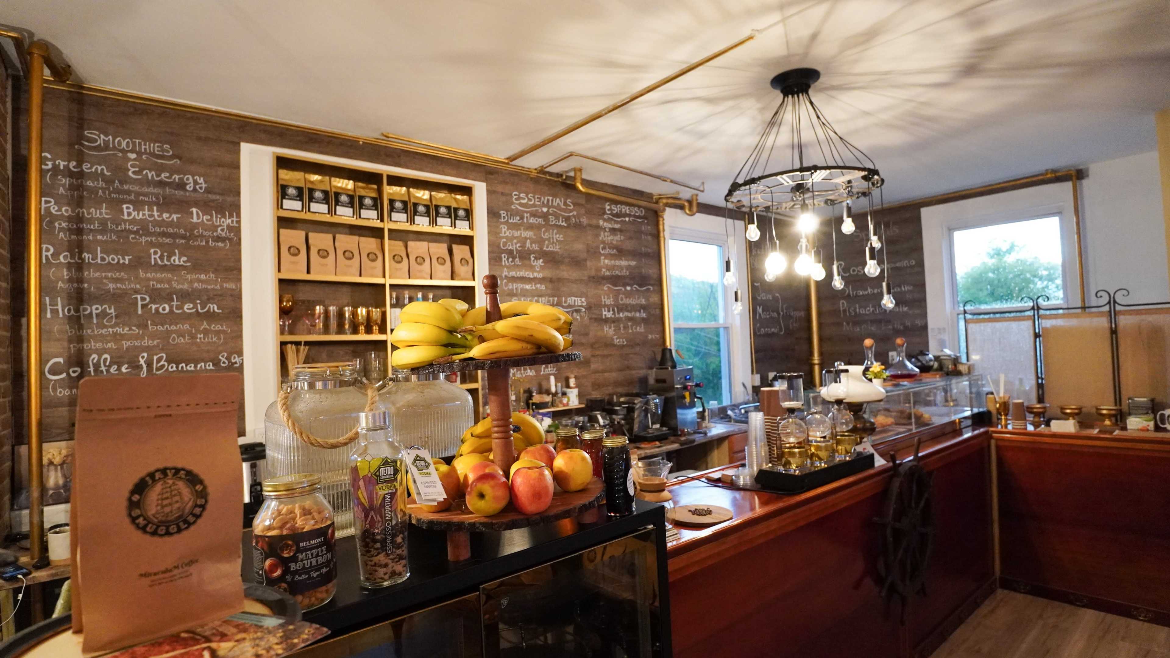 A cozy coffee shop interior with a wooden counter displaying assorted fruits and jars, chalkboard menus on the walls, and pendant lighting above.