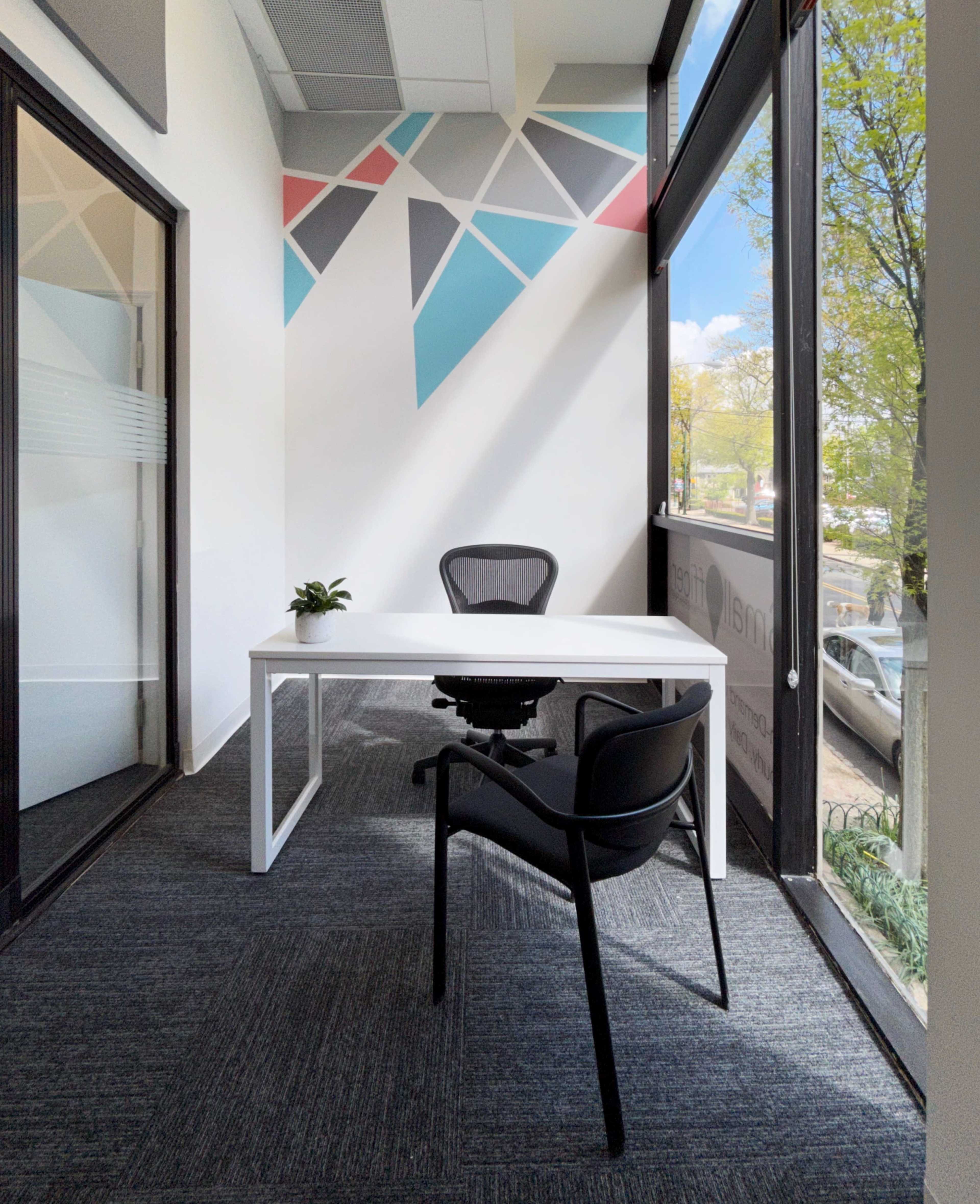 A modern office space features a white desk and a black chair against a wall with geometric designs and large windows.