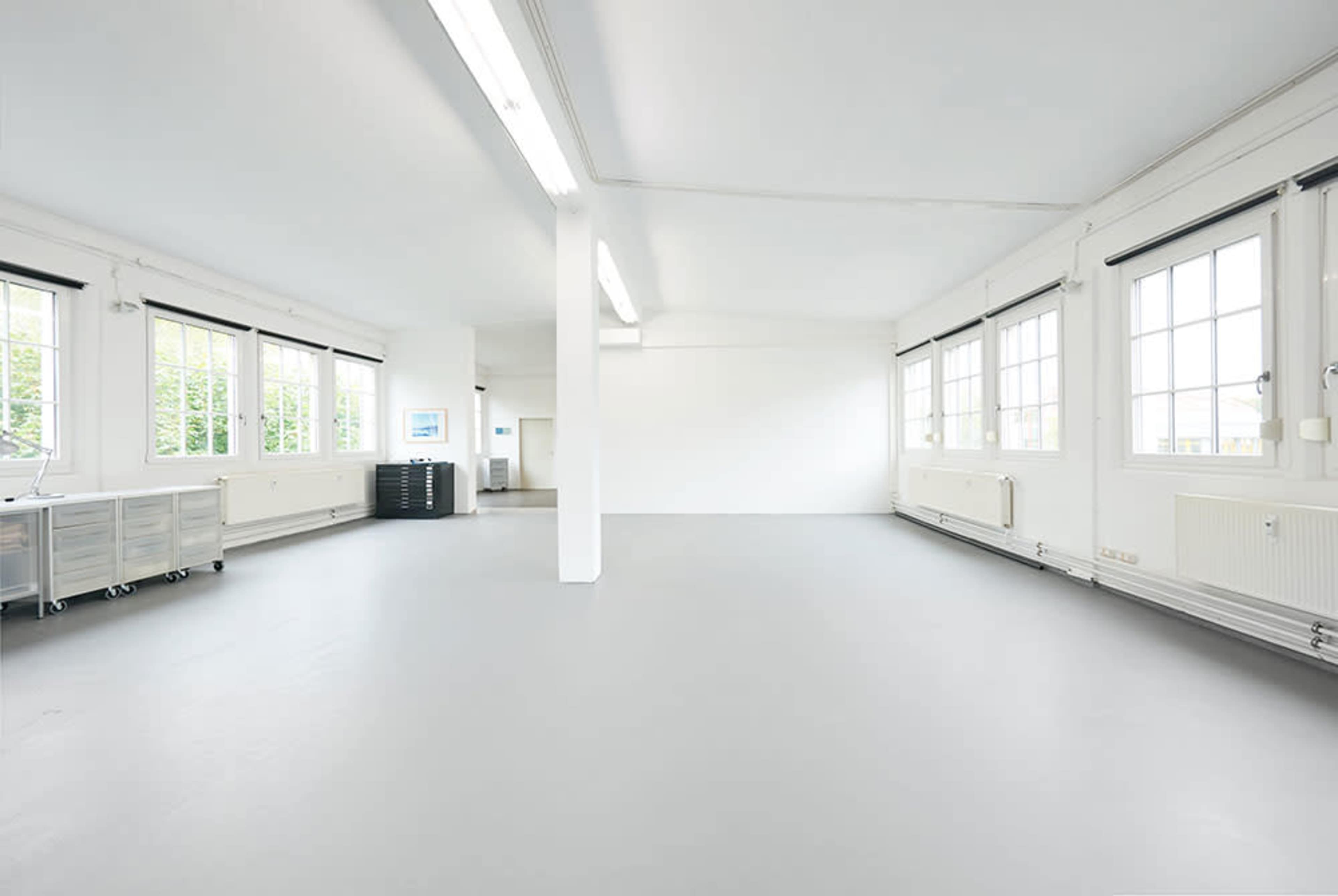 The image shows a spacious, empty room with large windows, white walls, and a gray floor.