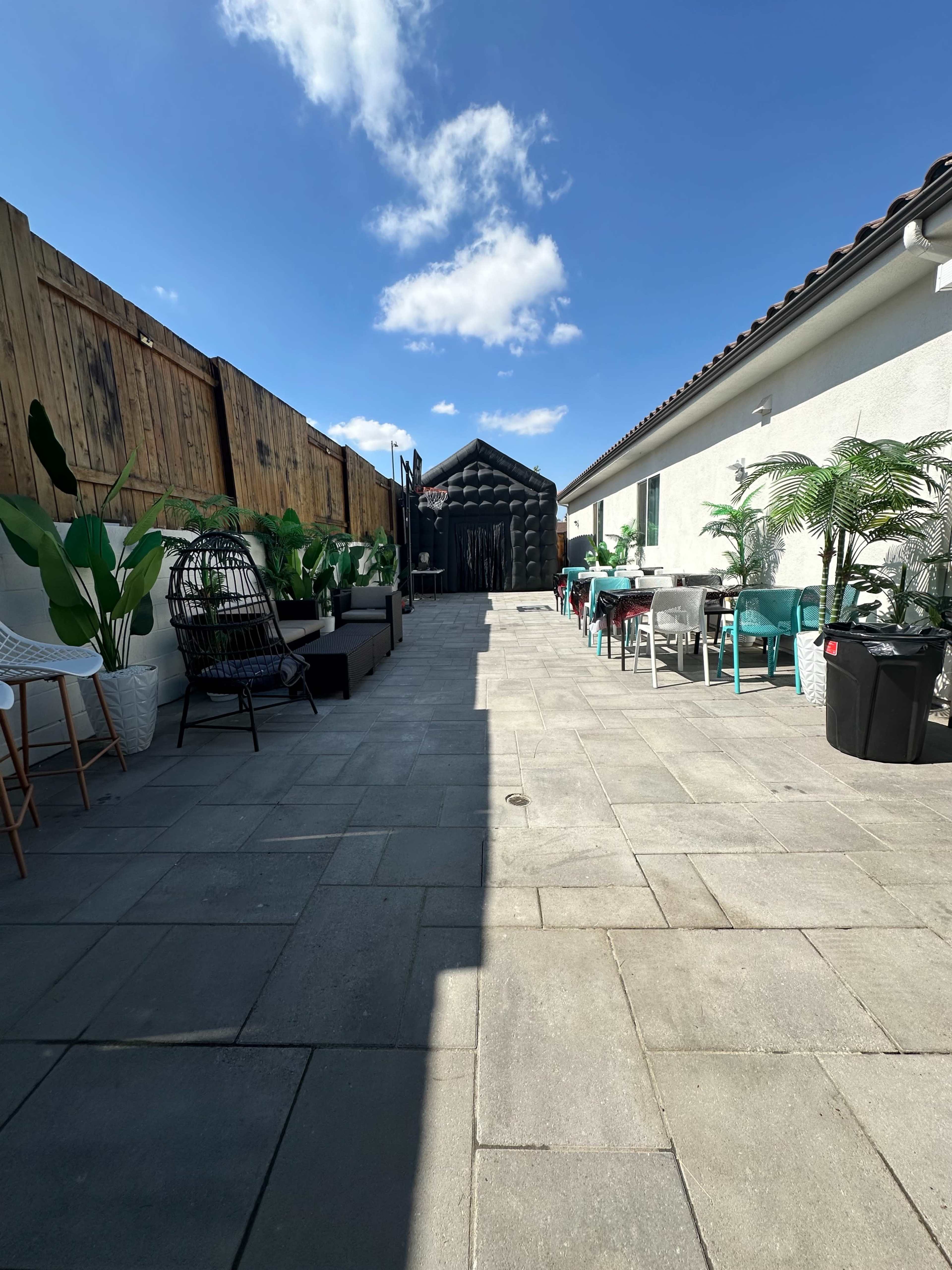 A narrow outdoor pathway lined with potted plants leads to a black structure at the end, flanked by seating areas and tables on either side.