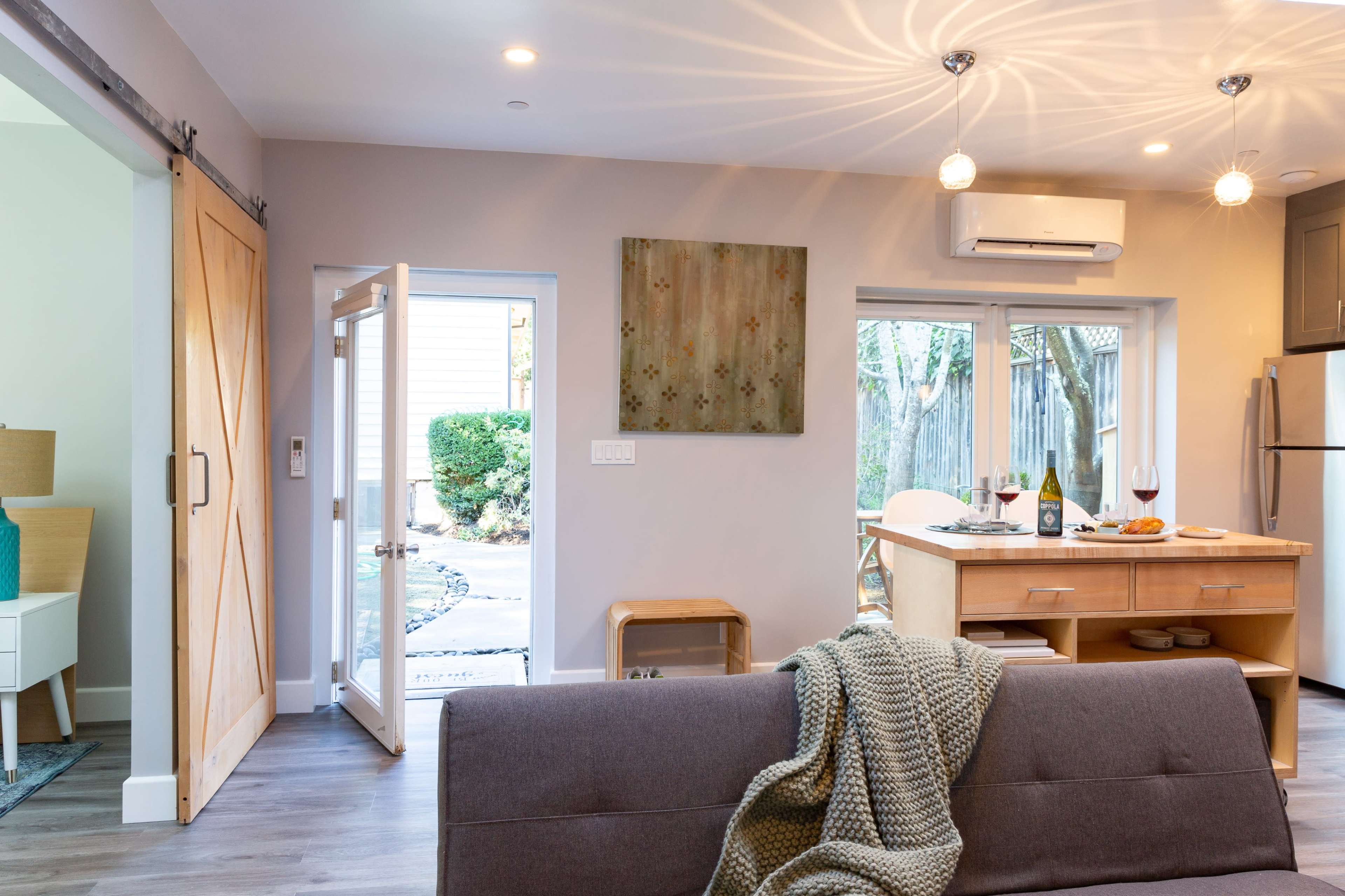 A cozy living space features a sofa, a wooden bar cart with wine and snacks, and a door leading to an outdoor area with greenery.