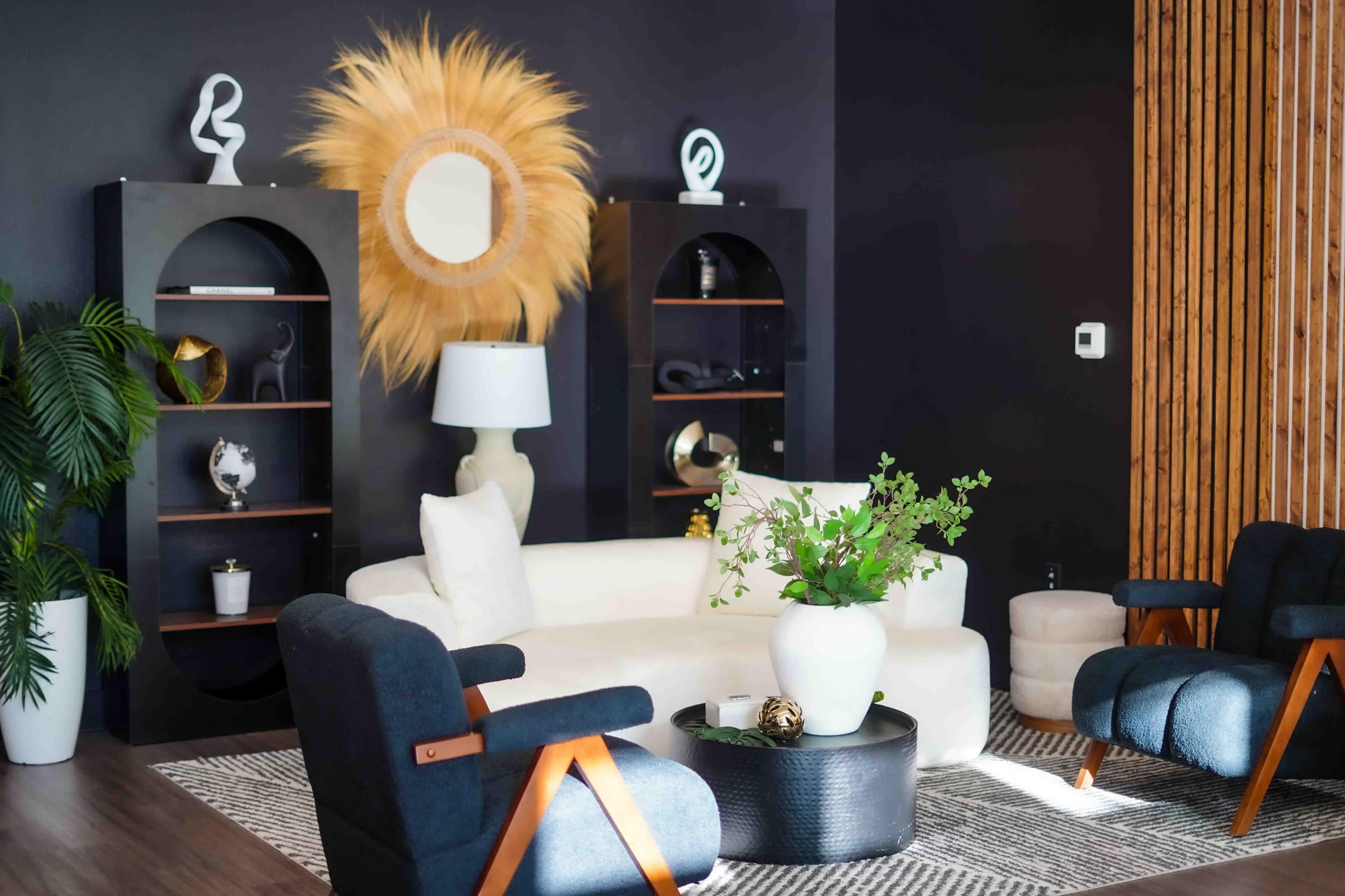 The image shows a modern living room featuring a white couch, black accent chairs, a round coffee table, and decorative shelving units against a dark wall.