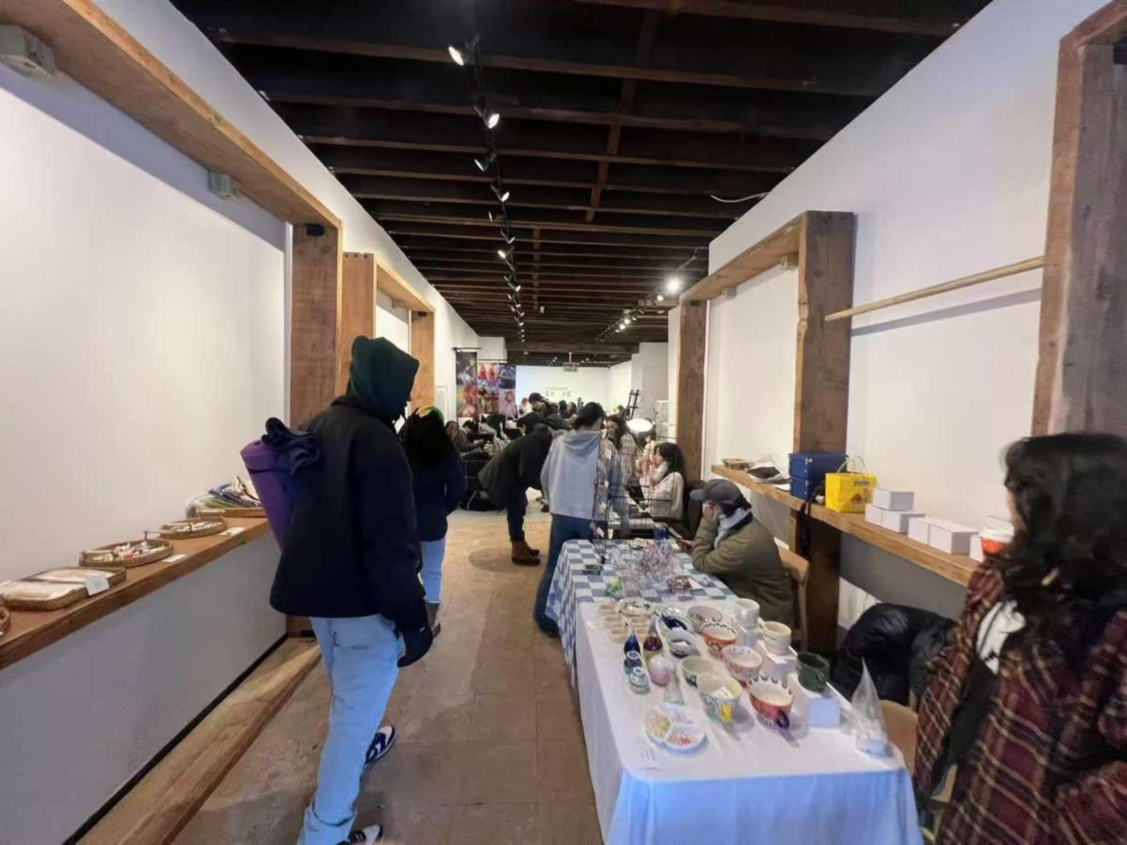 The image shows an industrial-style indoor market space with tables displaying various food items, while people browse and converse along the aisles.