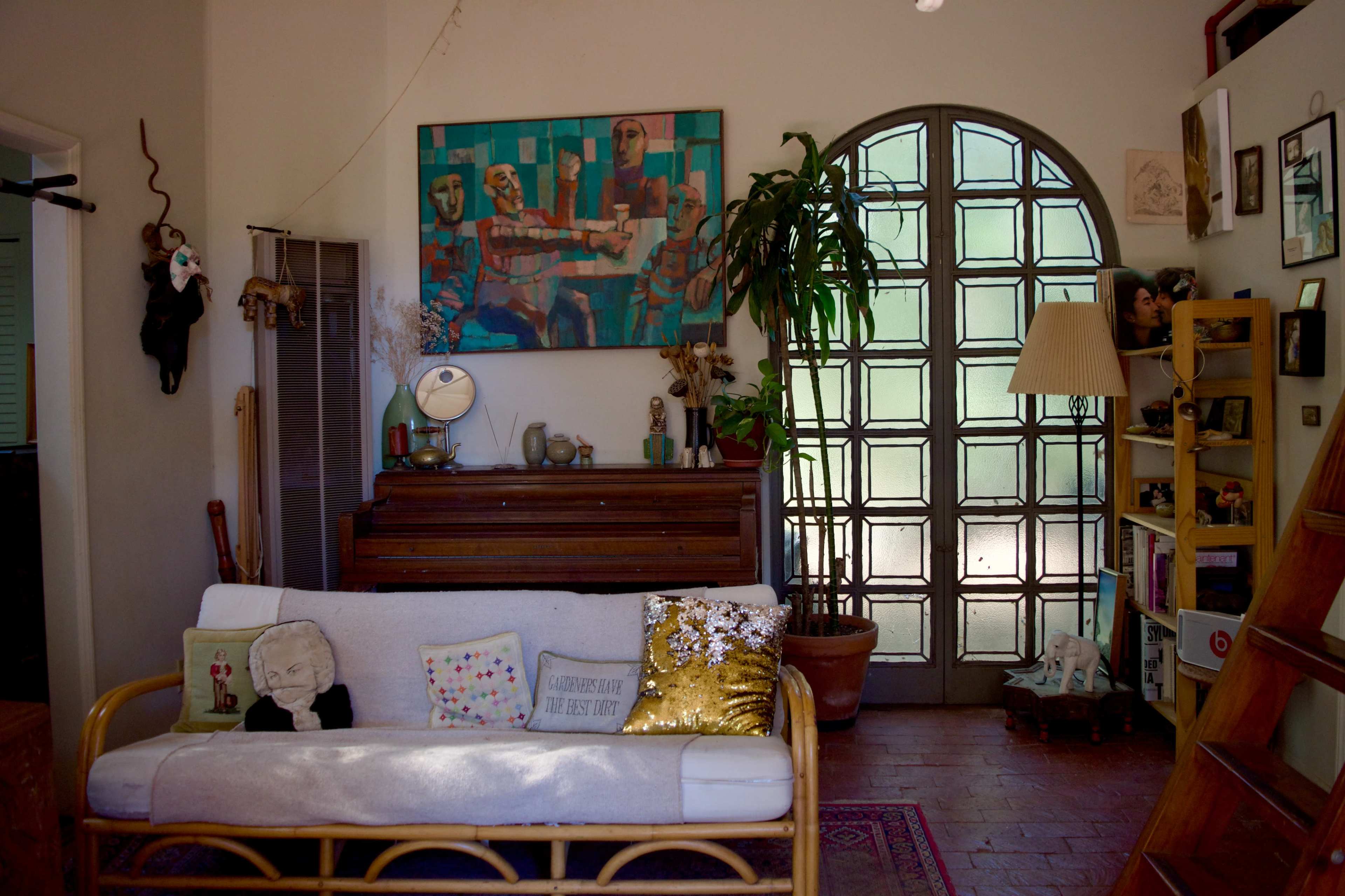 A cozy living room featuring a white sofa with decorative pillows, a wooden piano, and a large abstract painting on the wall, alongside a tall glass door and a collection of shelves filled with various items.