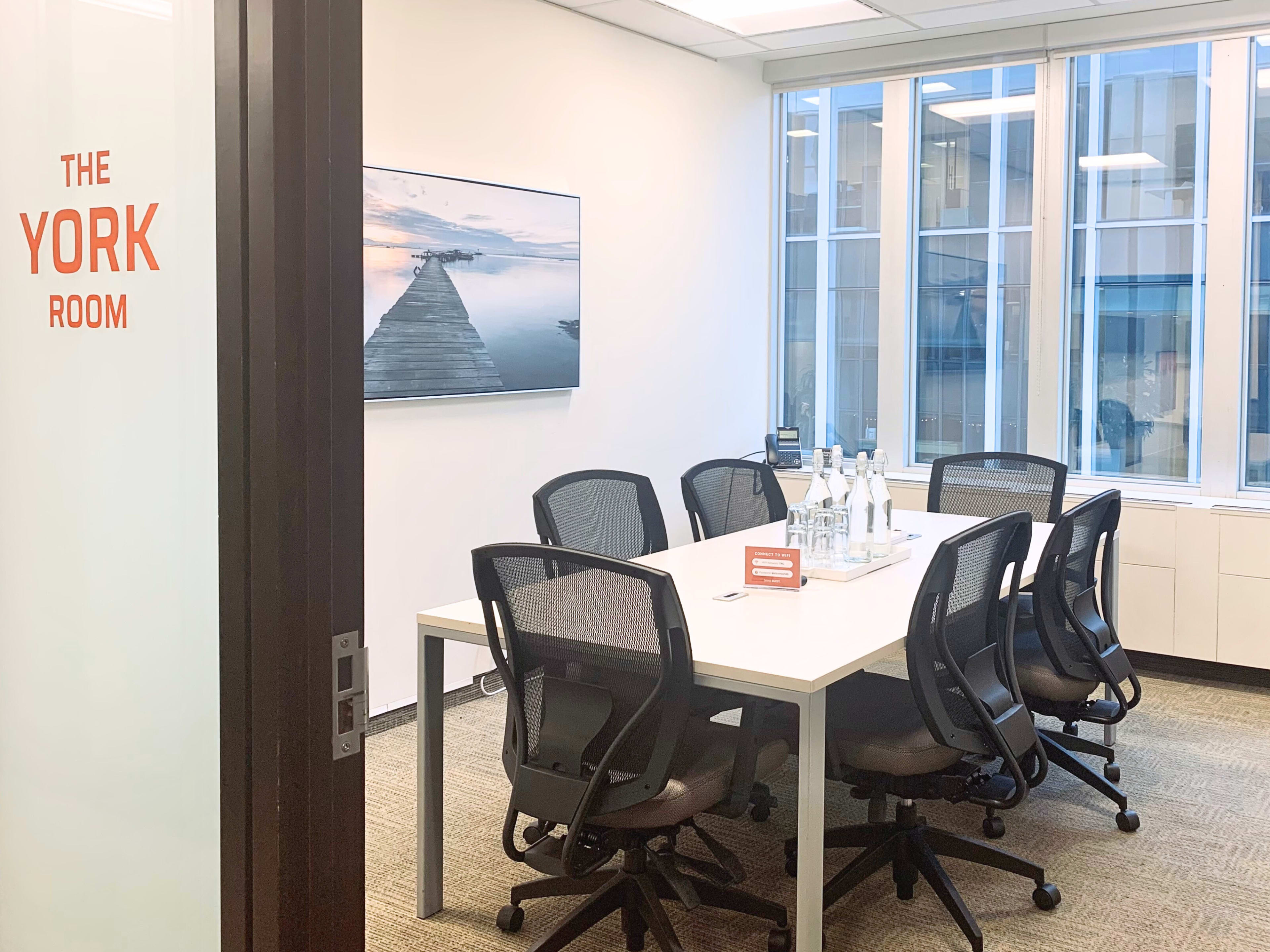 A conference room with a large white table surrounded by six chairs and a large screen displaying a waterfront image.