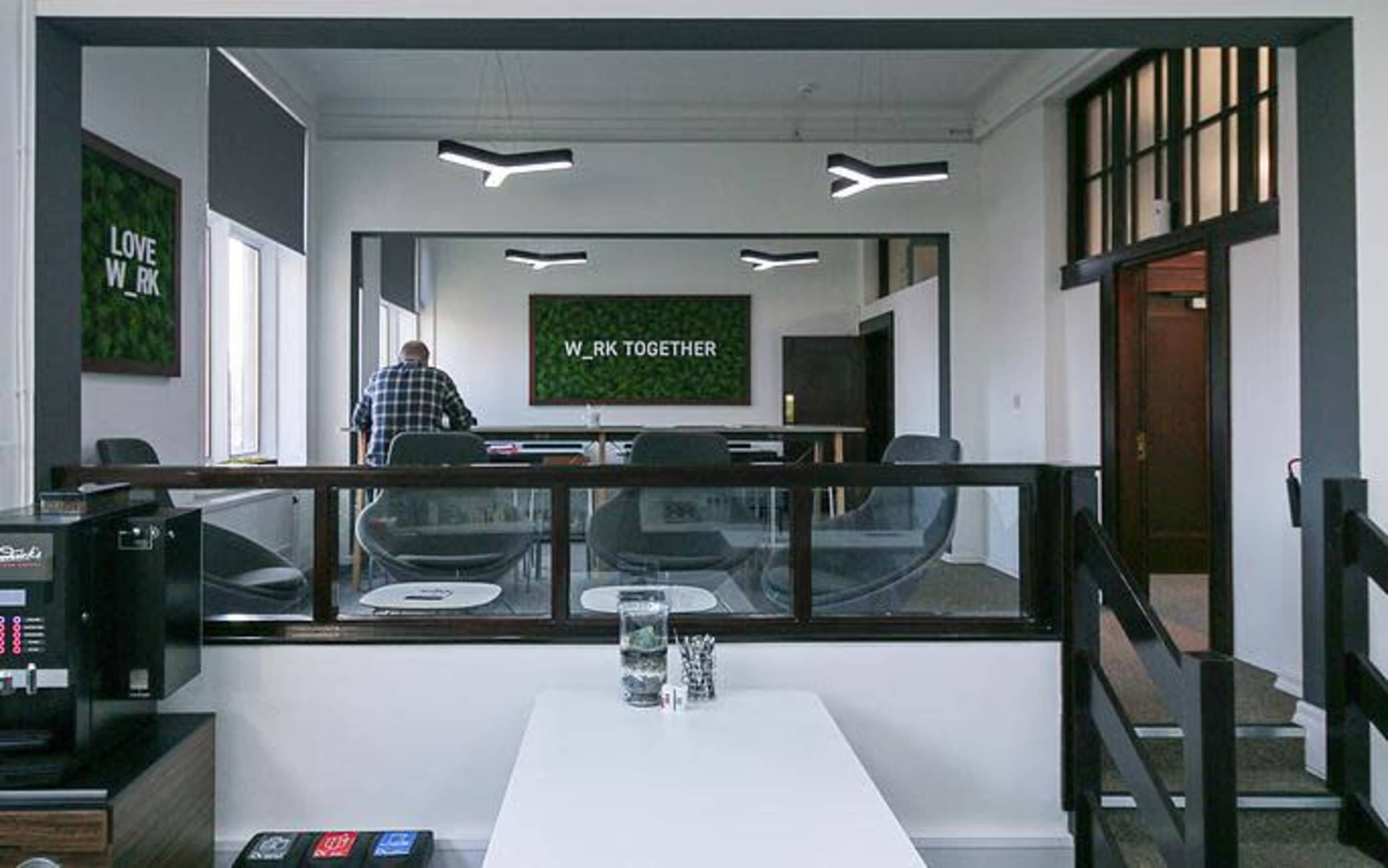 The image shows a modern office space with a meeting area visible in the background and a man standing near a desk in the foreground.
