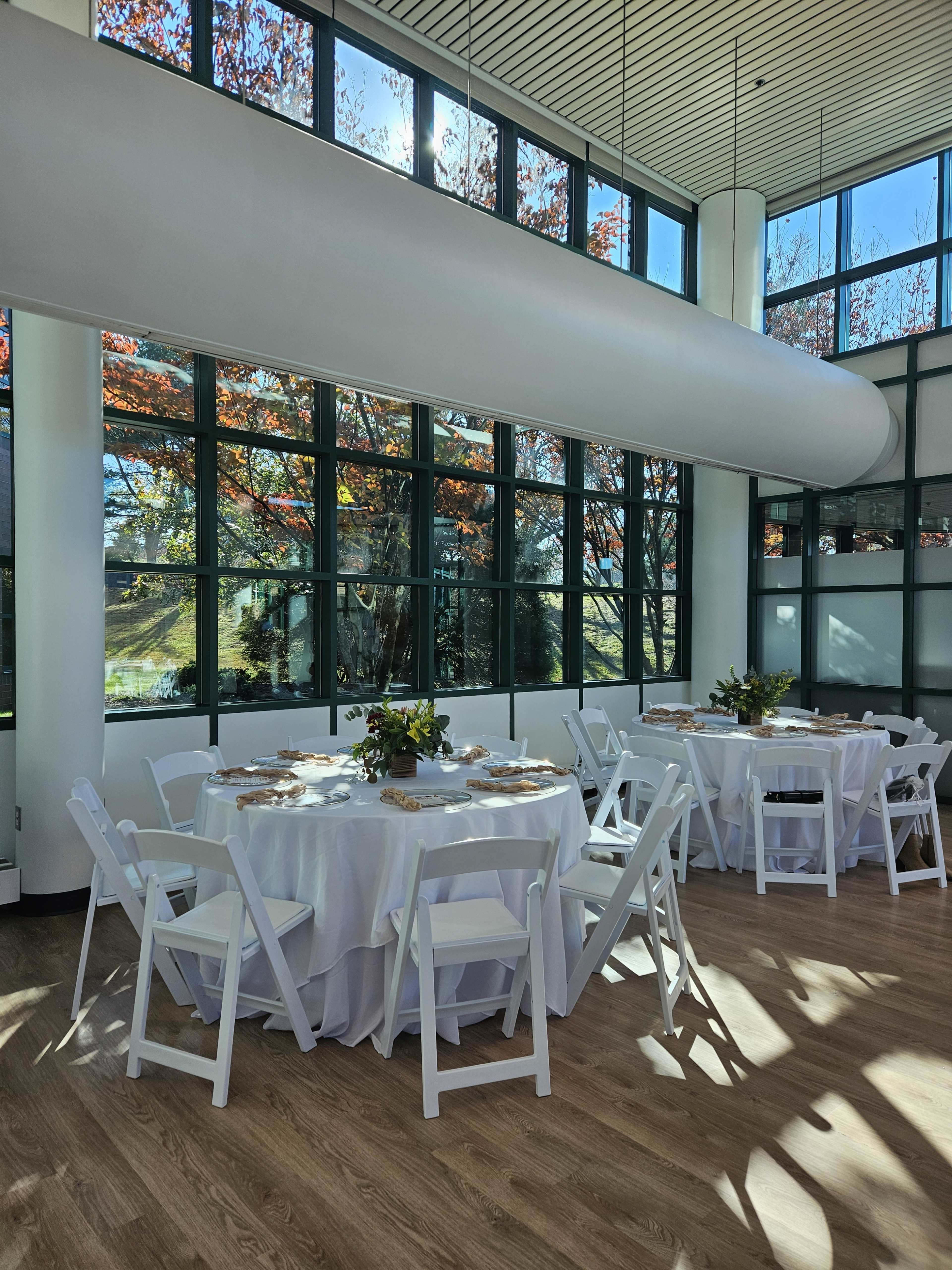 A modern dining space with white tables set for an event, surrounded by large windows displaying autumn foliage.