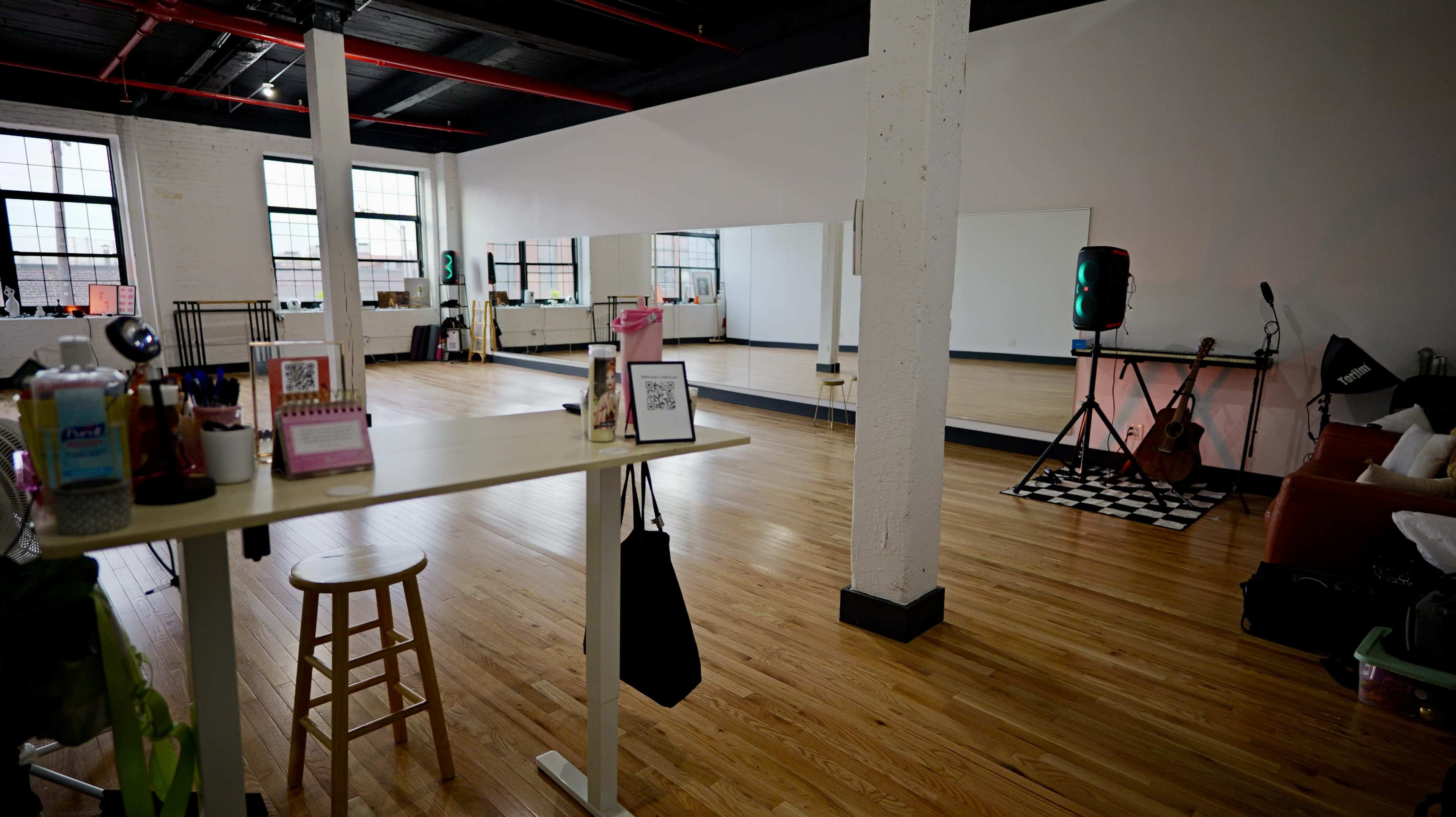The image shows a spacious, well-lit dance studio with wooden floors, mirrors, and a small stage setup at one end.