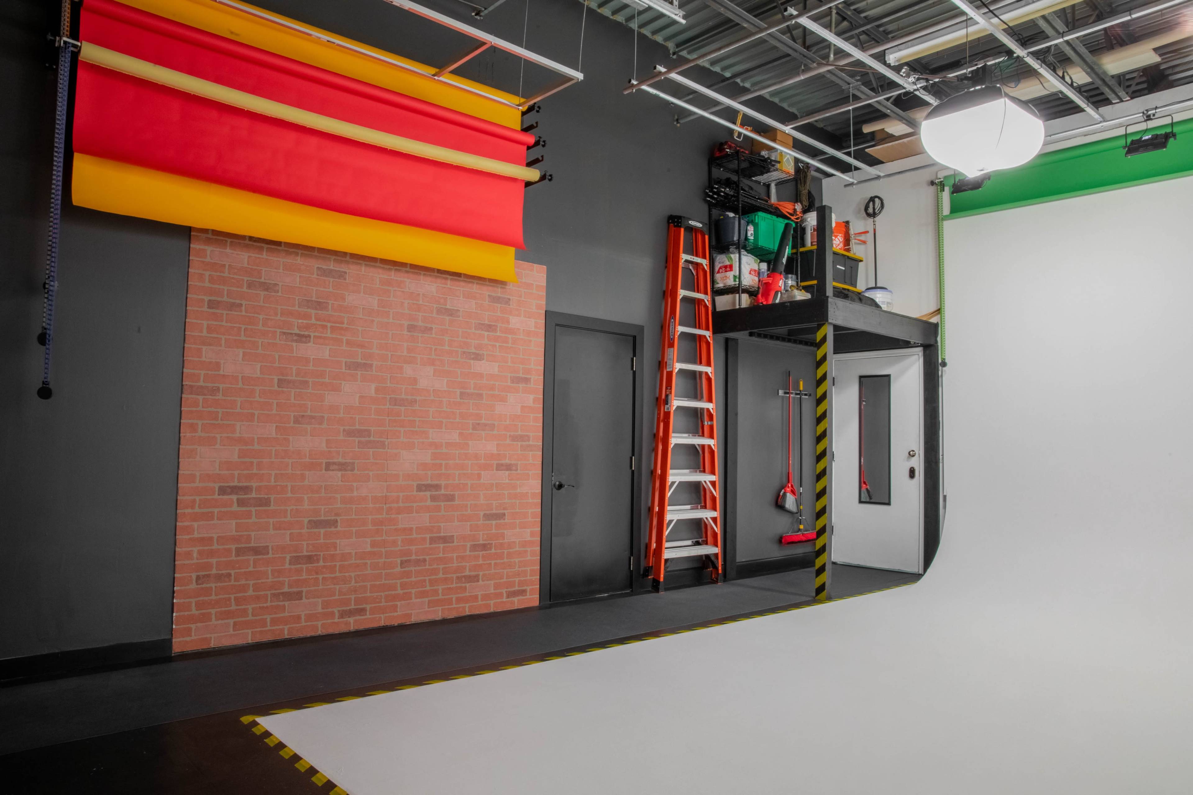 A photography studio with colorful backdrop rolls, a brick wall section, a ladder, and a clean white backdrop area.
