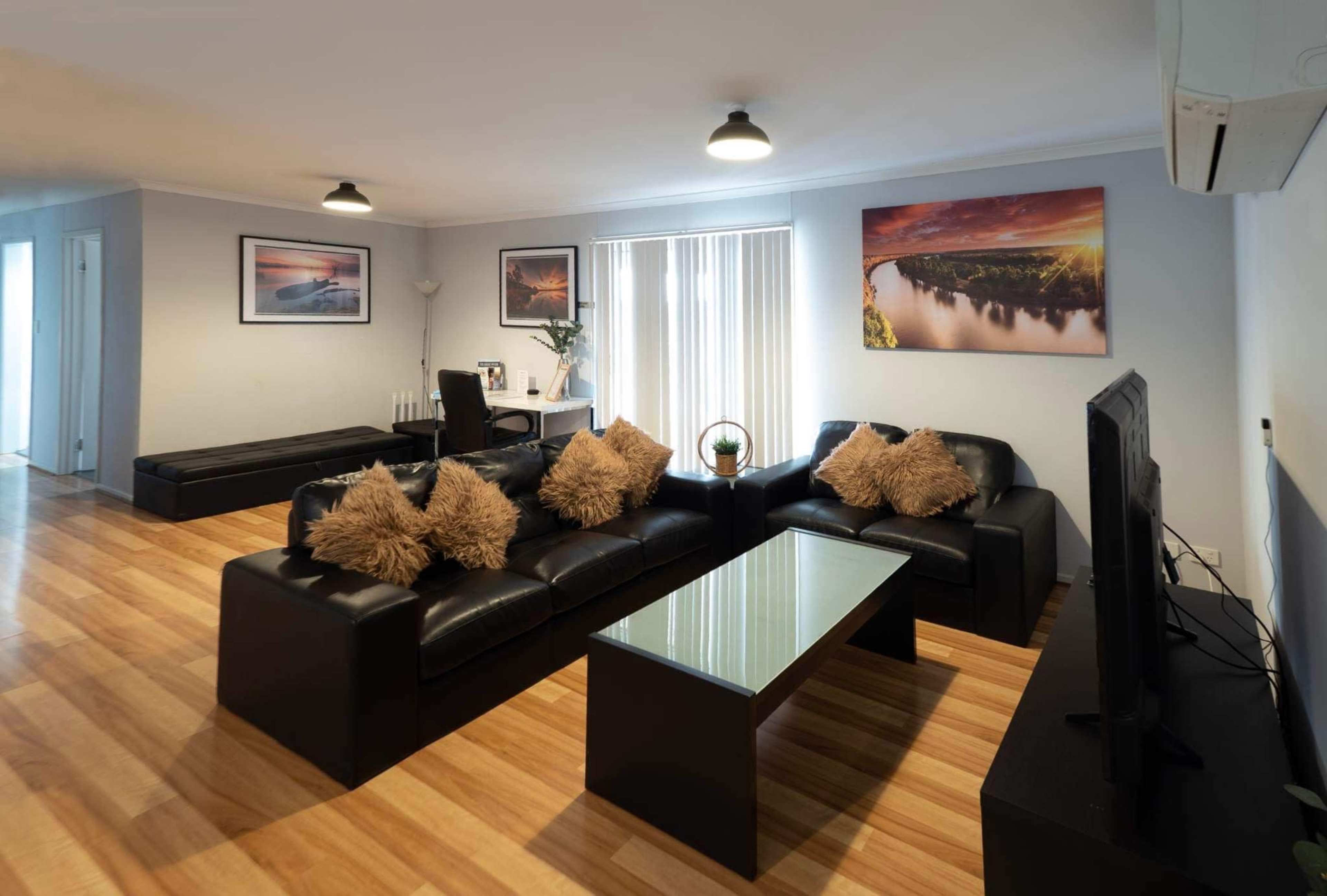 The image shows a modern living room featuring black leather sofas, a glass coffee table, and wooden flooring, with artwork on the walls and a television in the corner.