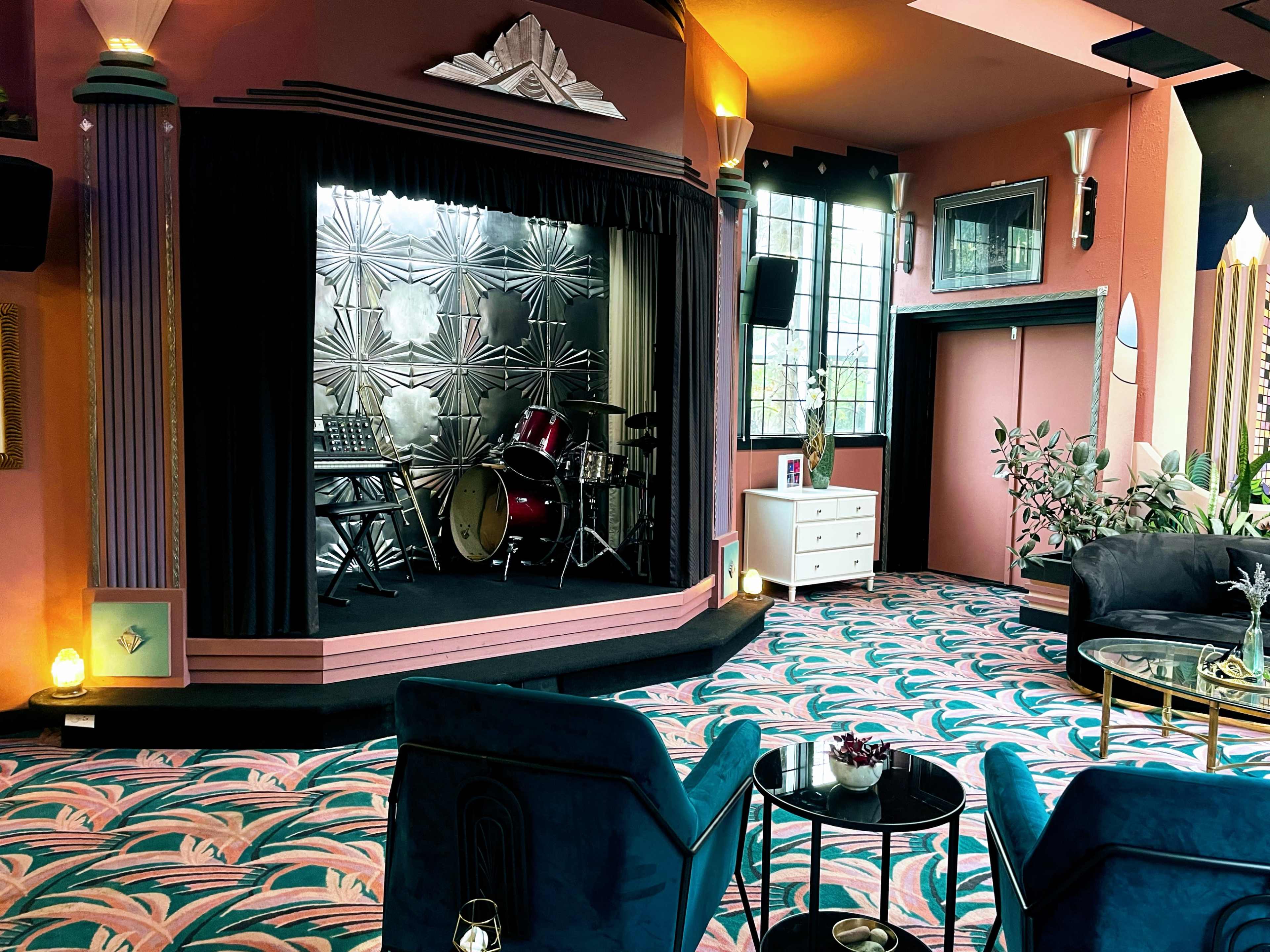 A stylish performance space featuring a band setup with drums and keyboards, surrounded by plush seating and vibrant patterned carpeting.