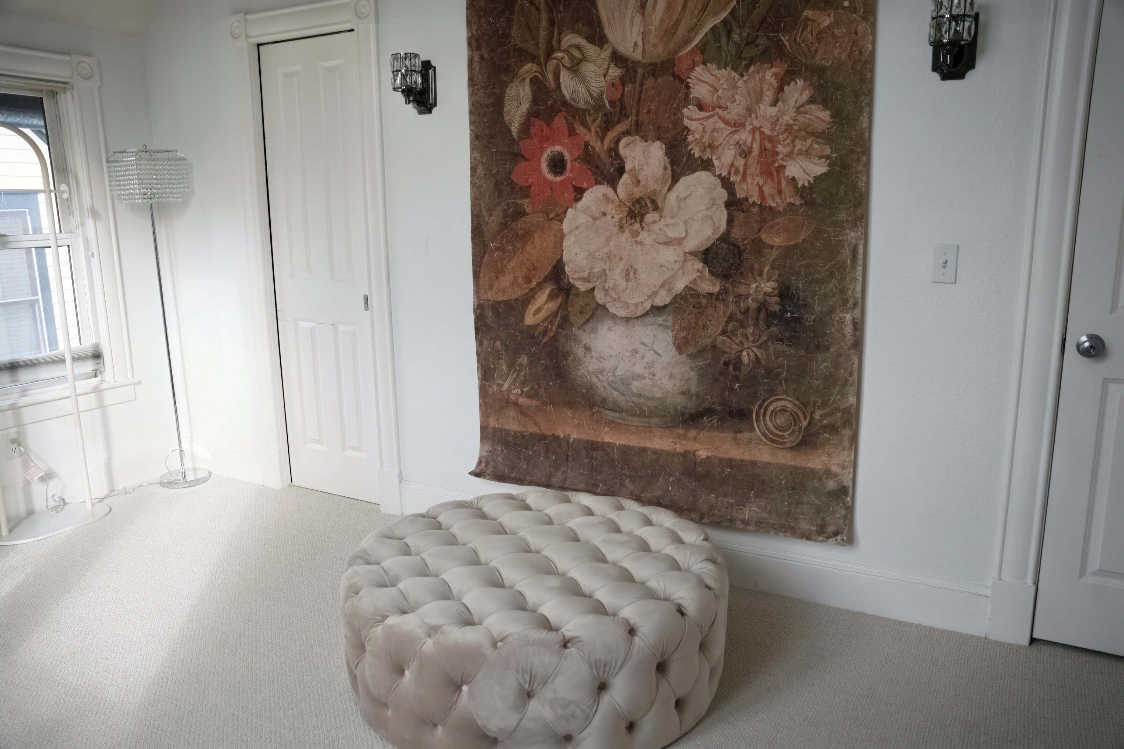 A large floral tapestry hangs on the wall above a round tufted ottoman in a softly lit room with white walls and a door.