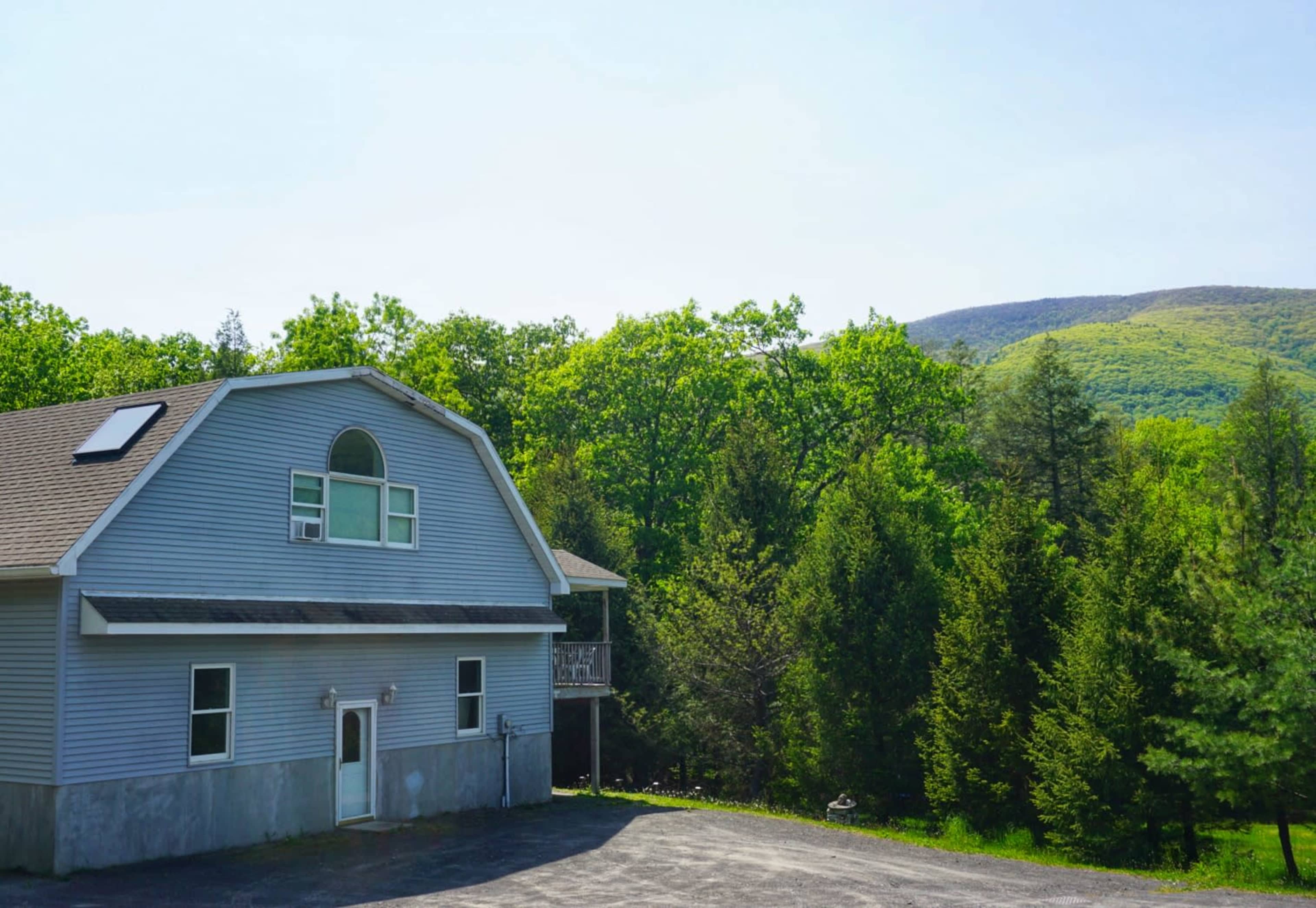 A gray house with a large front yard situated near a forested area and a mountain in the background under a clear sky.
