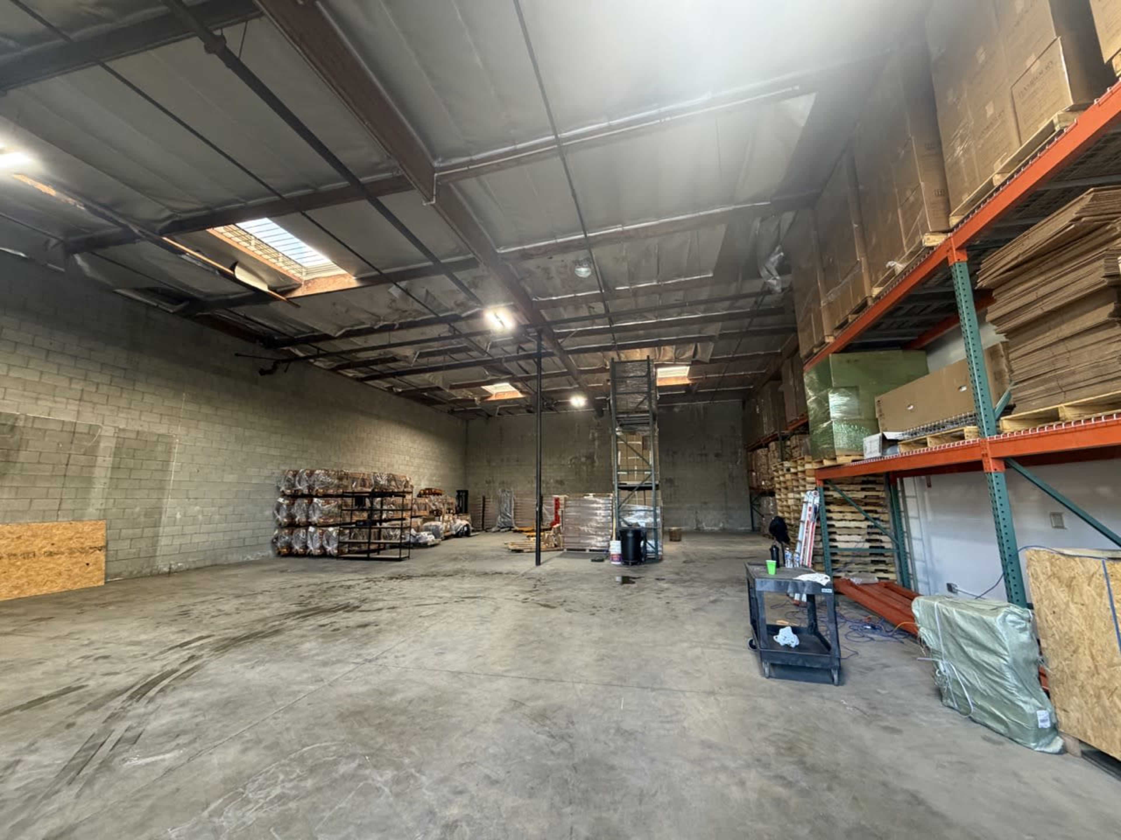 The image shows a large, empty warehouse space with shelves stocked on one side and pallets arranged on the floor.