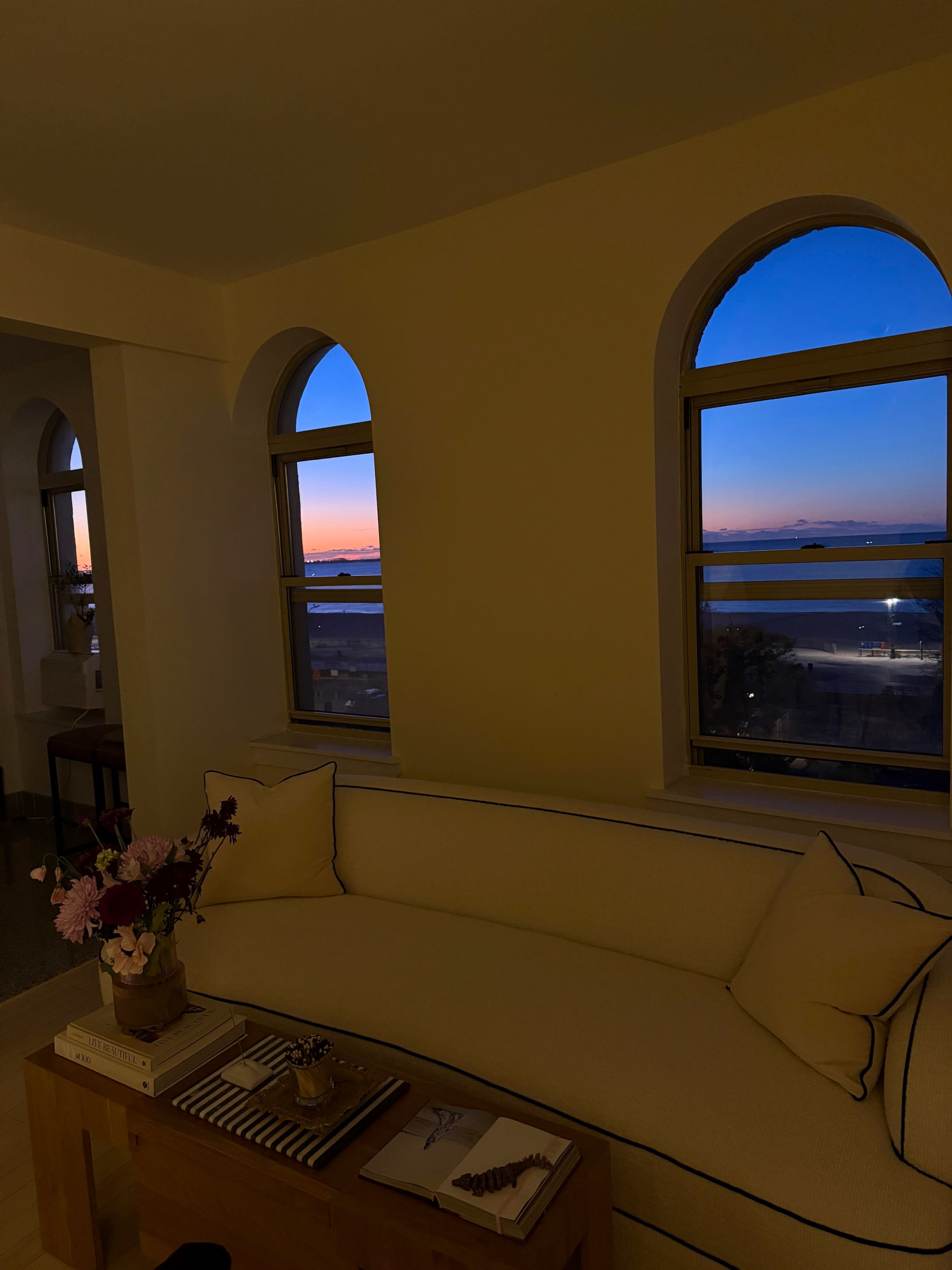 A cozy living room features a beige sofa and a coffee table, with large windows framing a view of a sunset over the water.