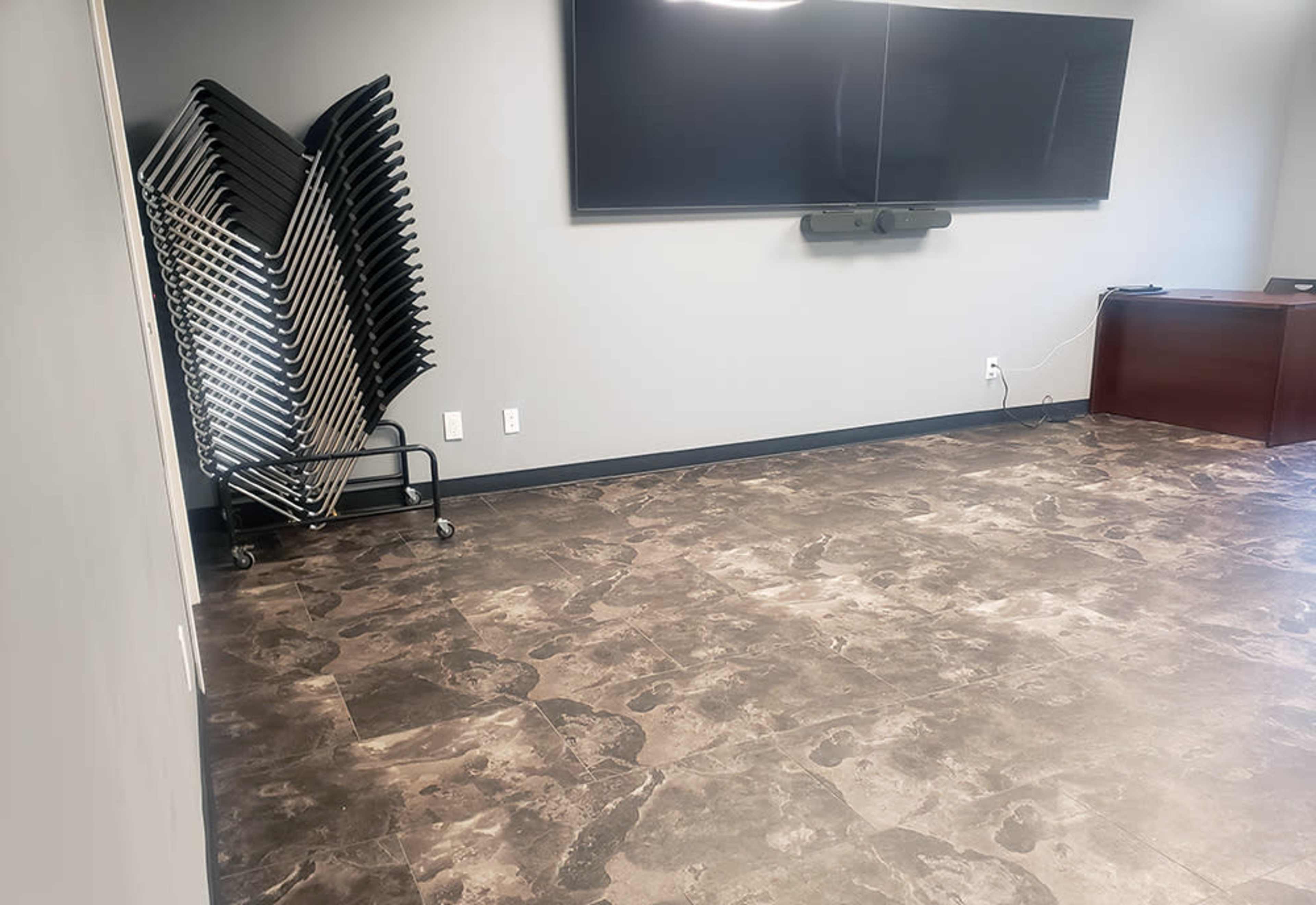 The image shows a sparsely furnished room with a large wall-mounted screen, a stack of chairs on a cart in the corner, and a dark wooden desk against the wall.