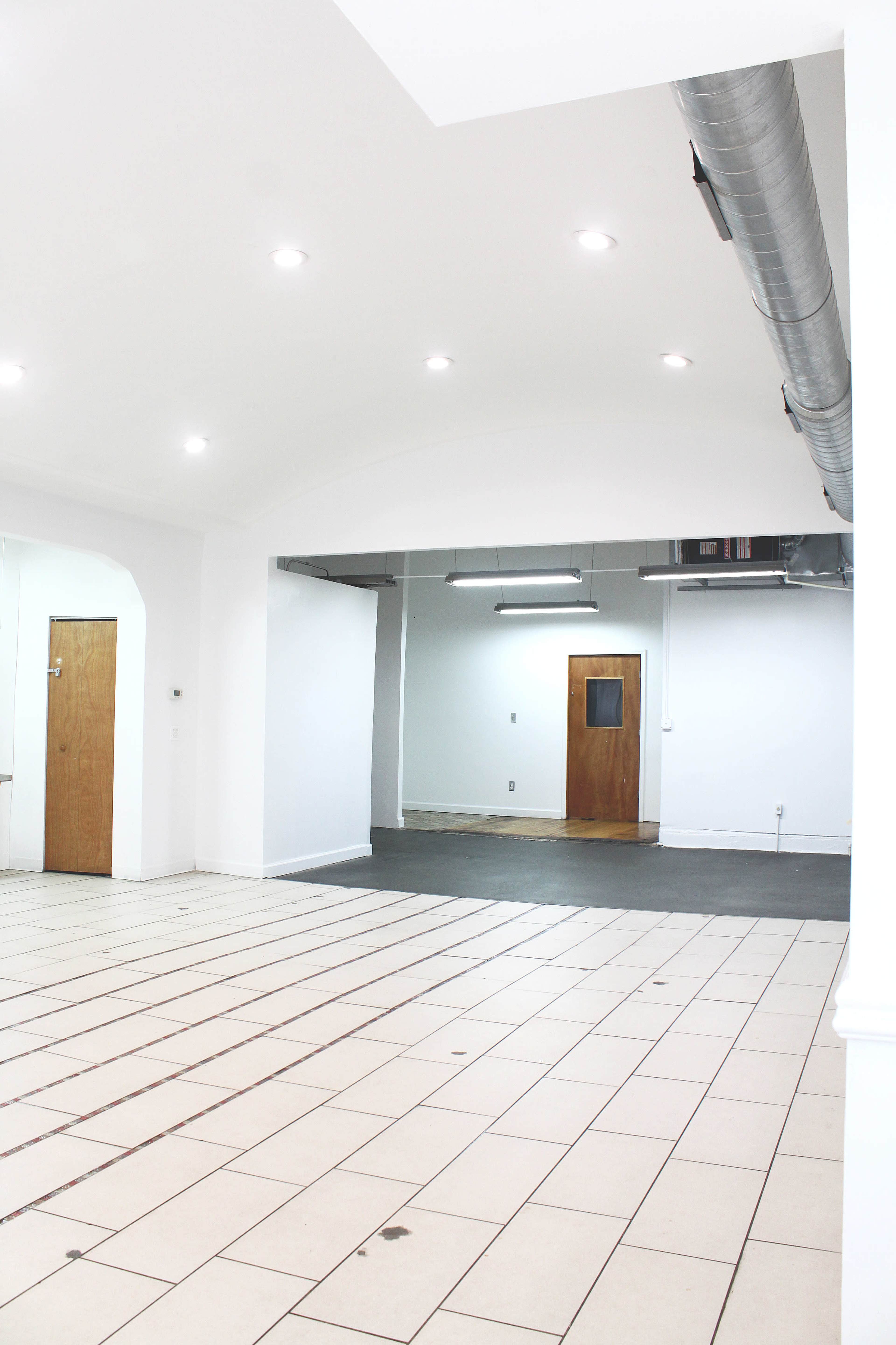 The image shows a spacious, empty room with white walls, tiled flooring, and a ceiling-mounted ventilation system.