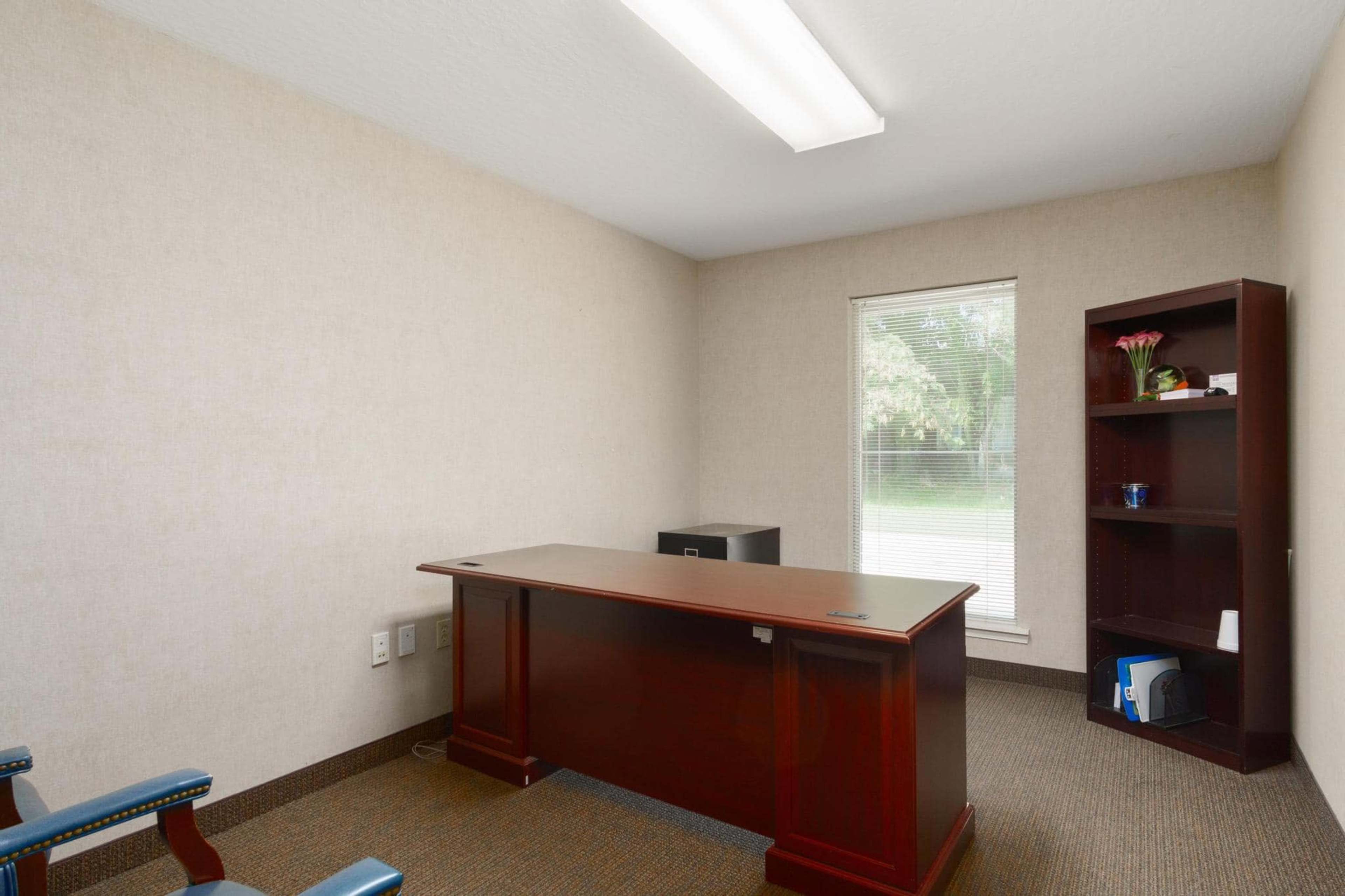 The image shows a small office with a wooden desk, a chair, a bookshelf, and a window allowing natural light.