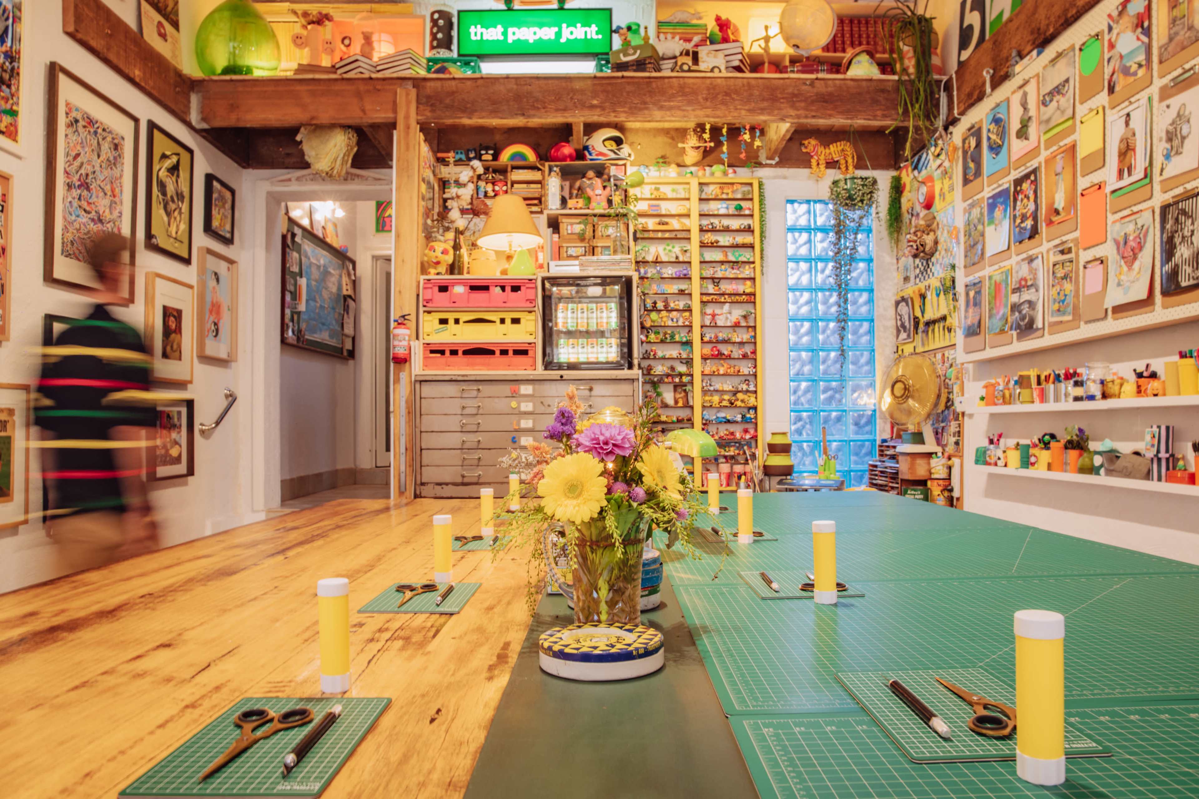 A colorful creative workspace filled with art supplies, a long table set for a project, and walls adorned with various framed artworks.