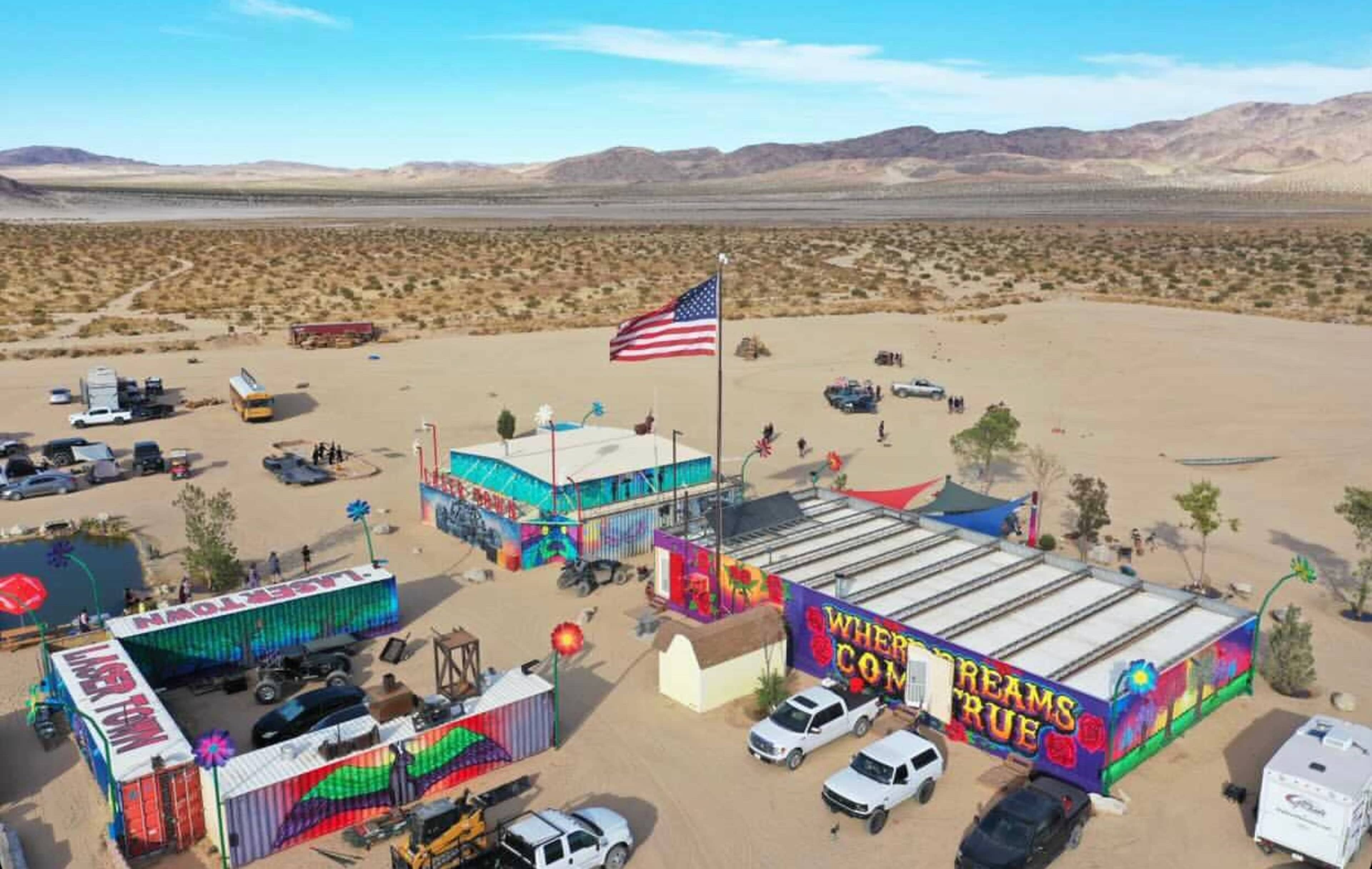 An aerial view shows a vibrant gathering in a desert landscape, featuring colorful structures, vehicles, and an American flag.