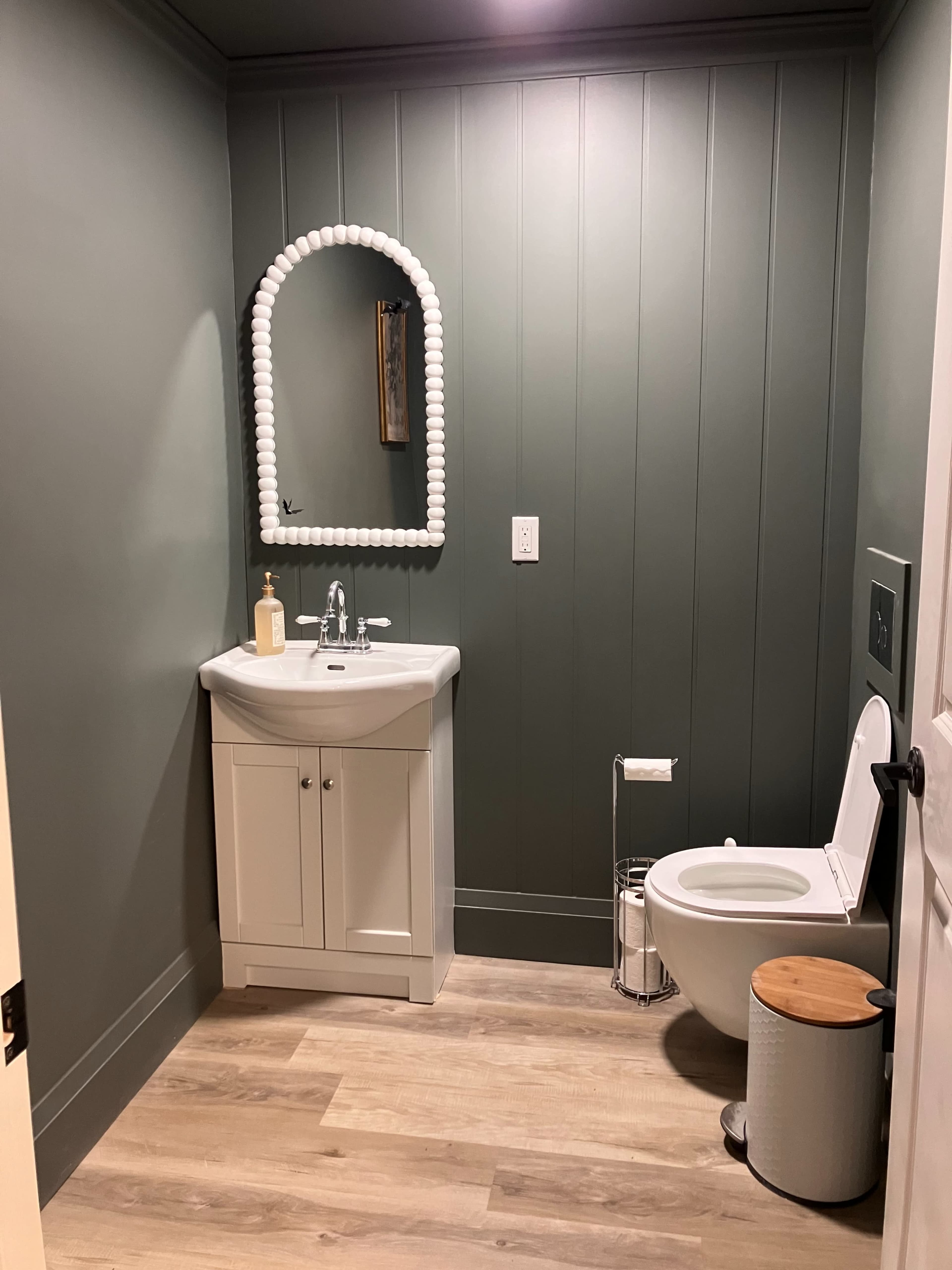 The image shows a small bathroom featuring a white vanity with a sink, a toilet, and a round, decorative mirror on a green paneled wall.