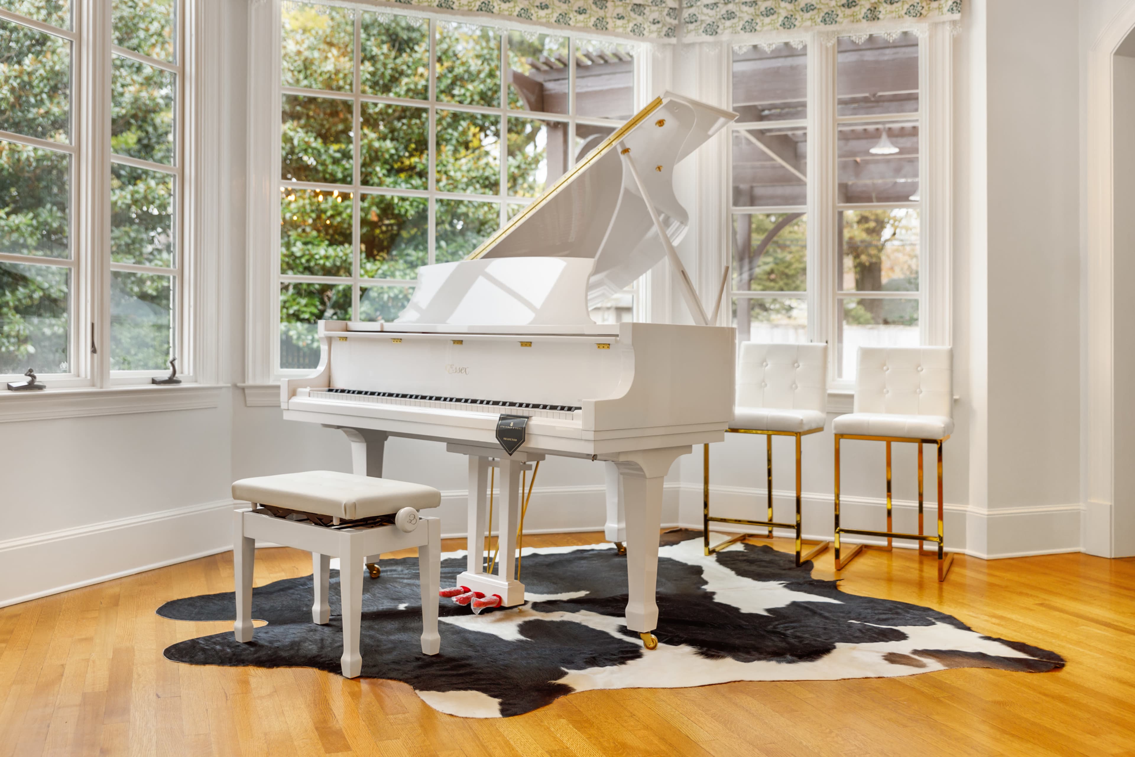 A white grand piano sits on a cowhide rug beside two bar stools in a bright room with large windows.