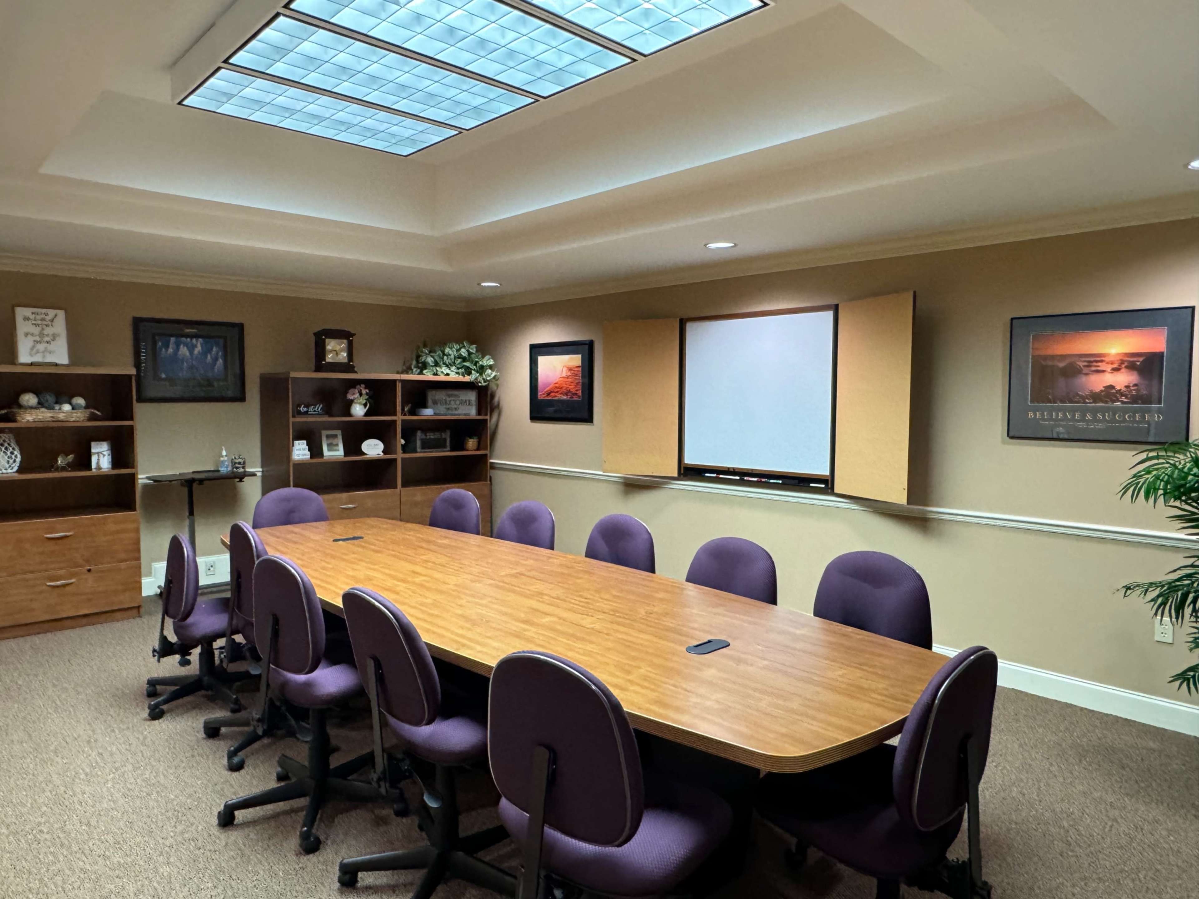A conference room features a long wooden table surrounded by purple chairs, with a projector screen and windows above for natural light.