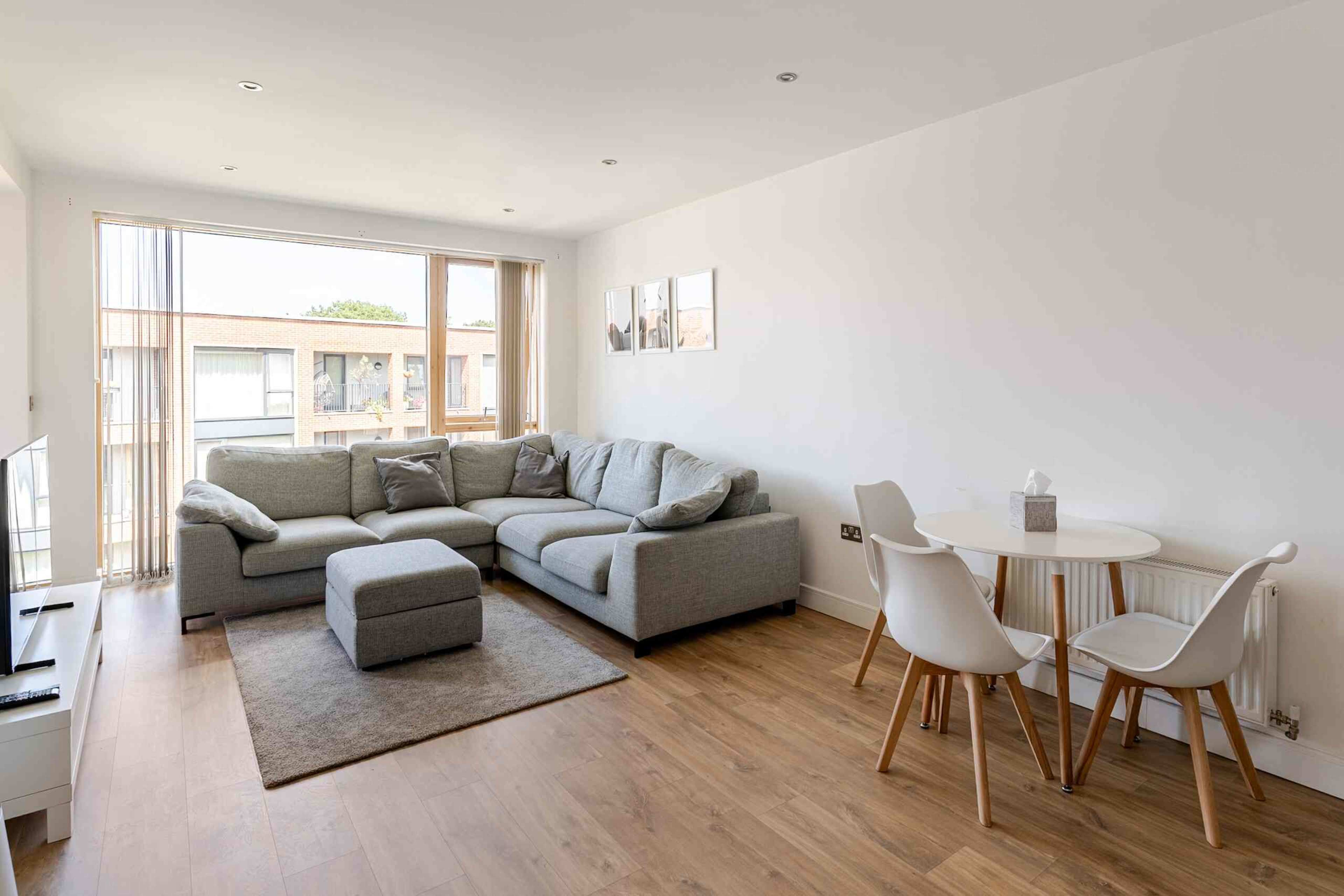 The image shows a modern, bright living room featuring a grey sectional sofa, a small round dining table with white chairs, and a large window allowing natural light to enter.