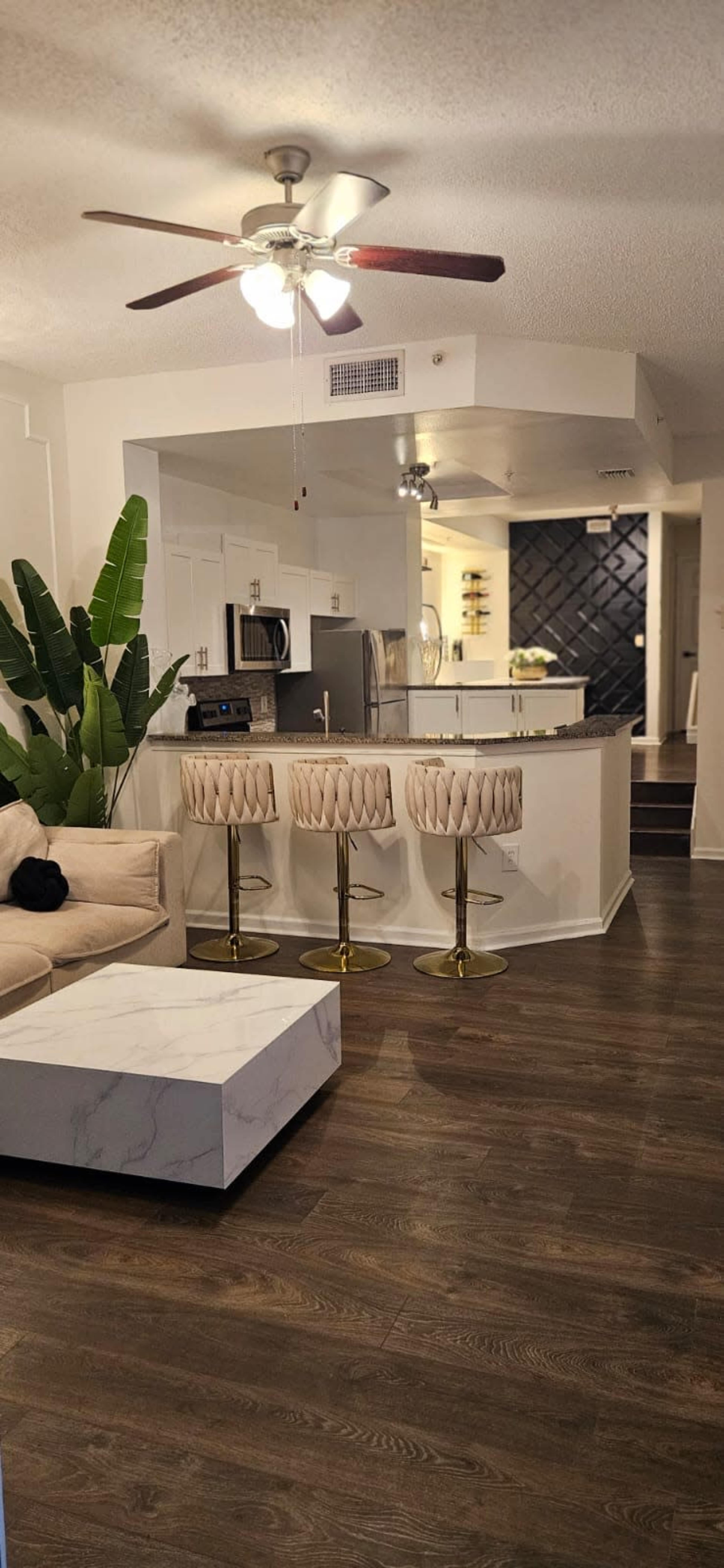 The image shows a modern living area with a light-colored sofa, three bar stools at a counter, a dining table, and a kitchen in the background, featuring a black accent wall.
