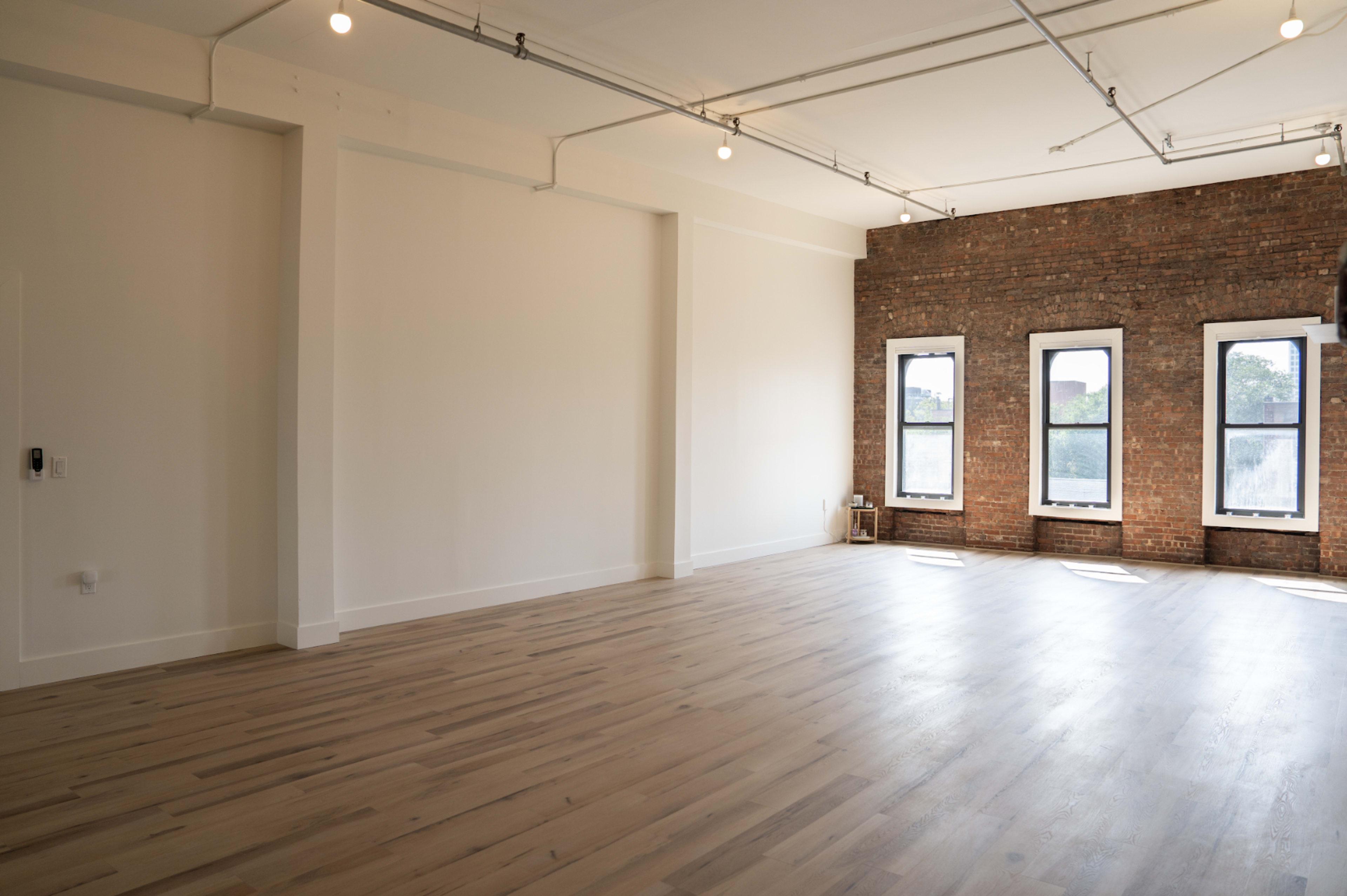 The image shows a spacious room with wooden flooring, exposed brick walls, and large windows allowing natural light to enter.