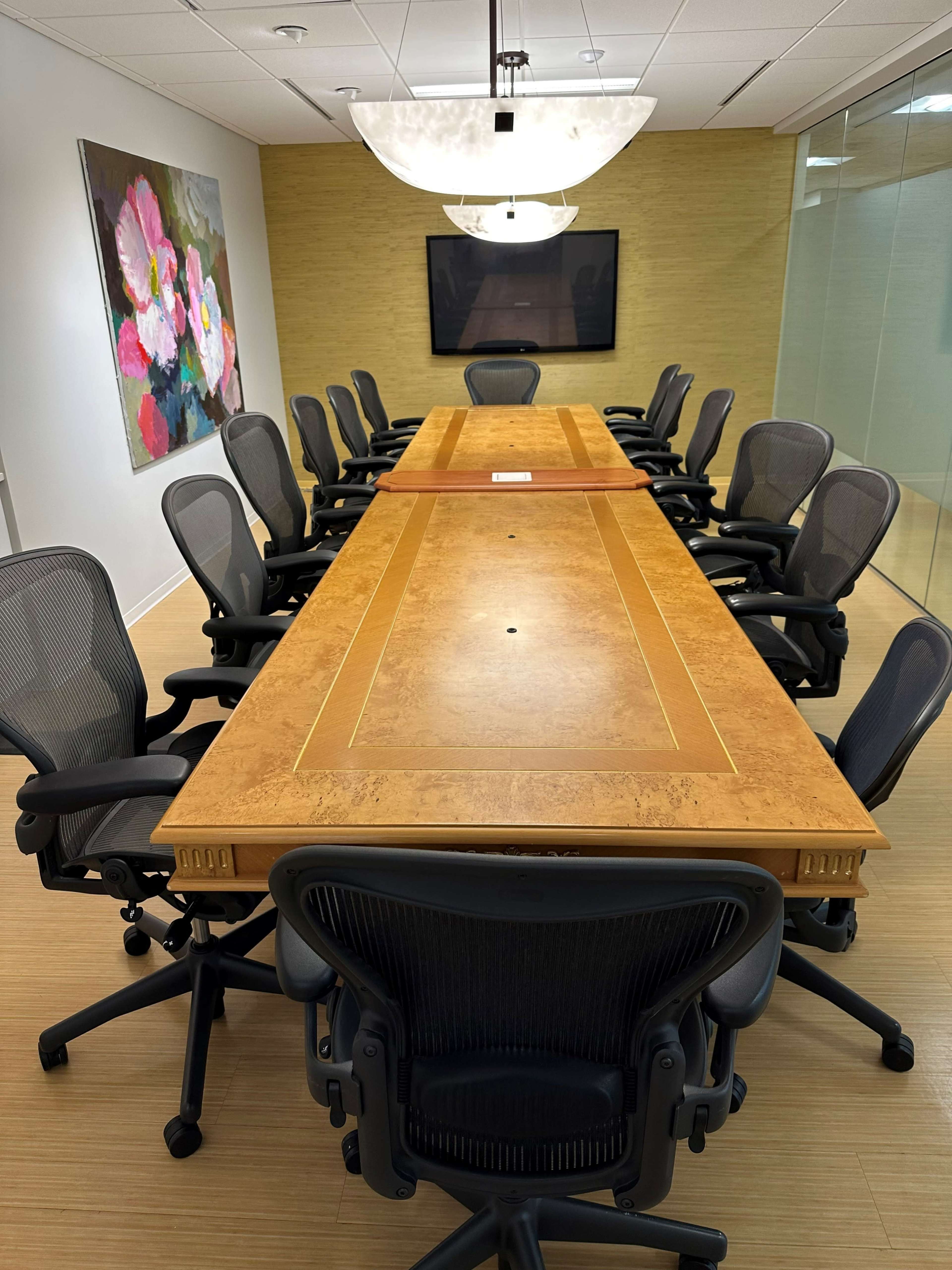 A long wooden conference table surrounded by ergonomic chairs is situated in a well-lit meeting room with a large screen and floral artwork on the wall.