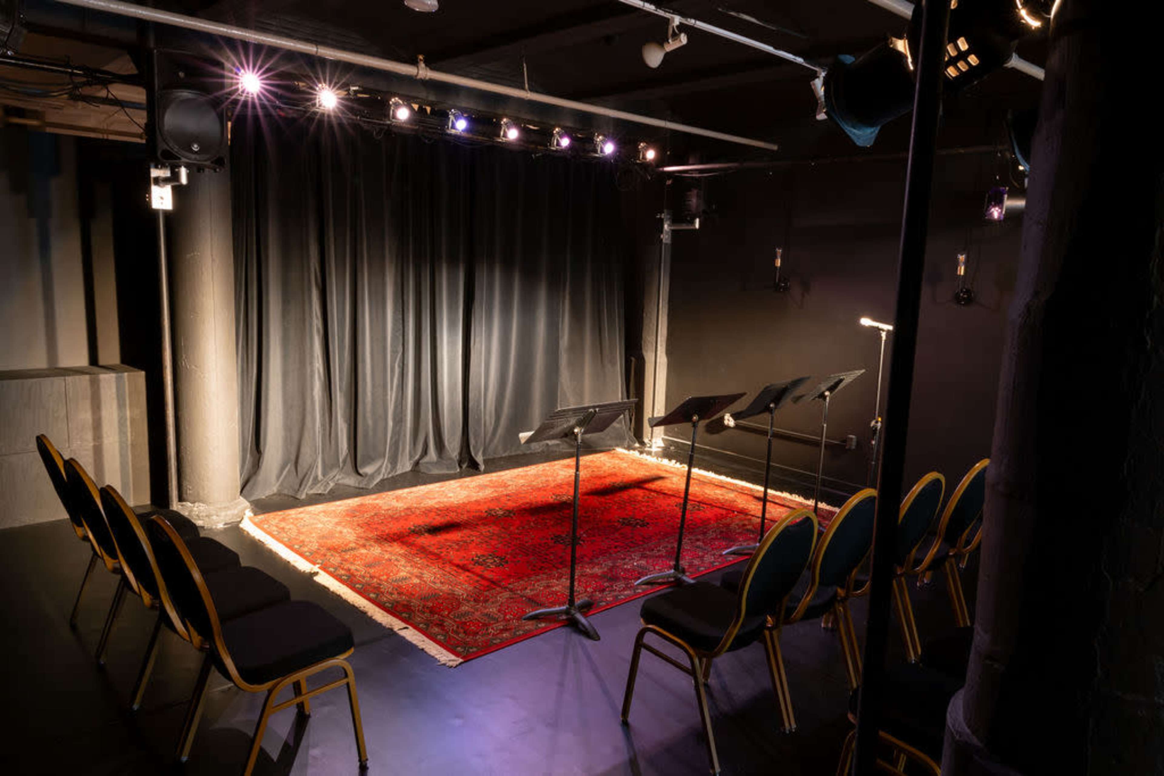 A small theater stage set with a red rug, black curtains, and music stands arranged in front of empty chairs.