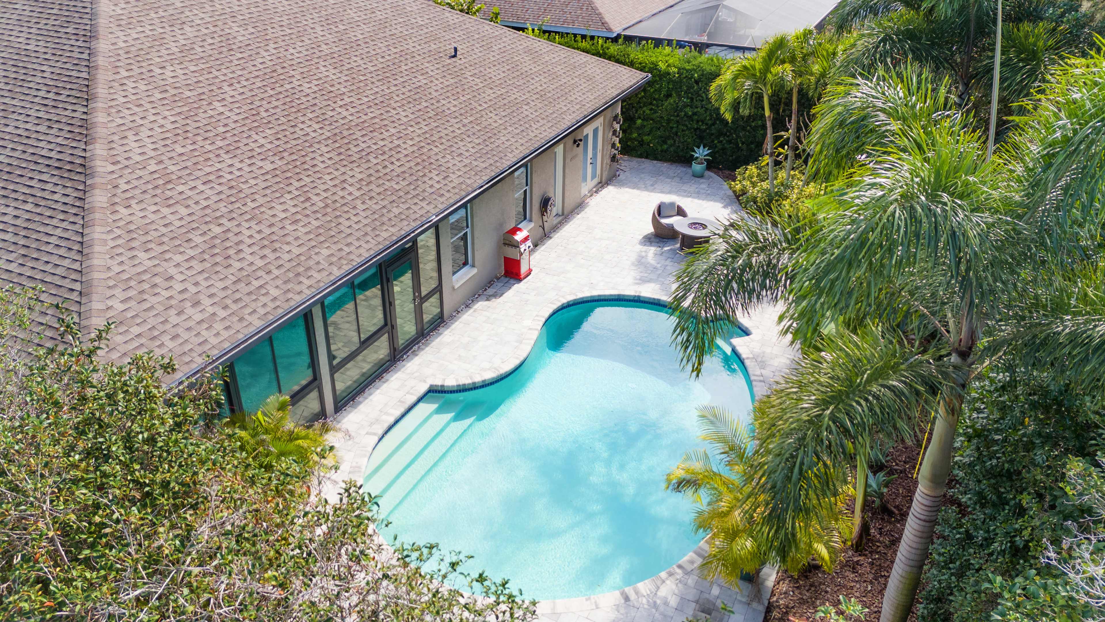 A backyard features a kidney-shaped pool surrounded by a patio and lush greenery, with a house positioned nearby.