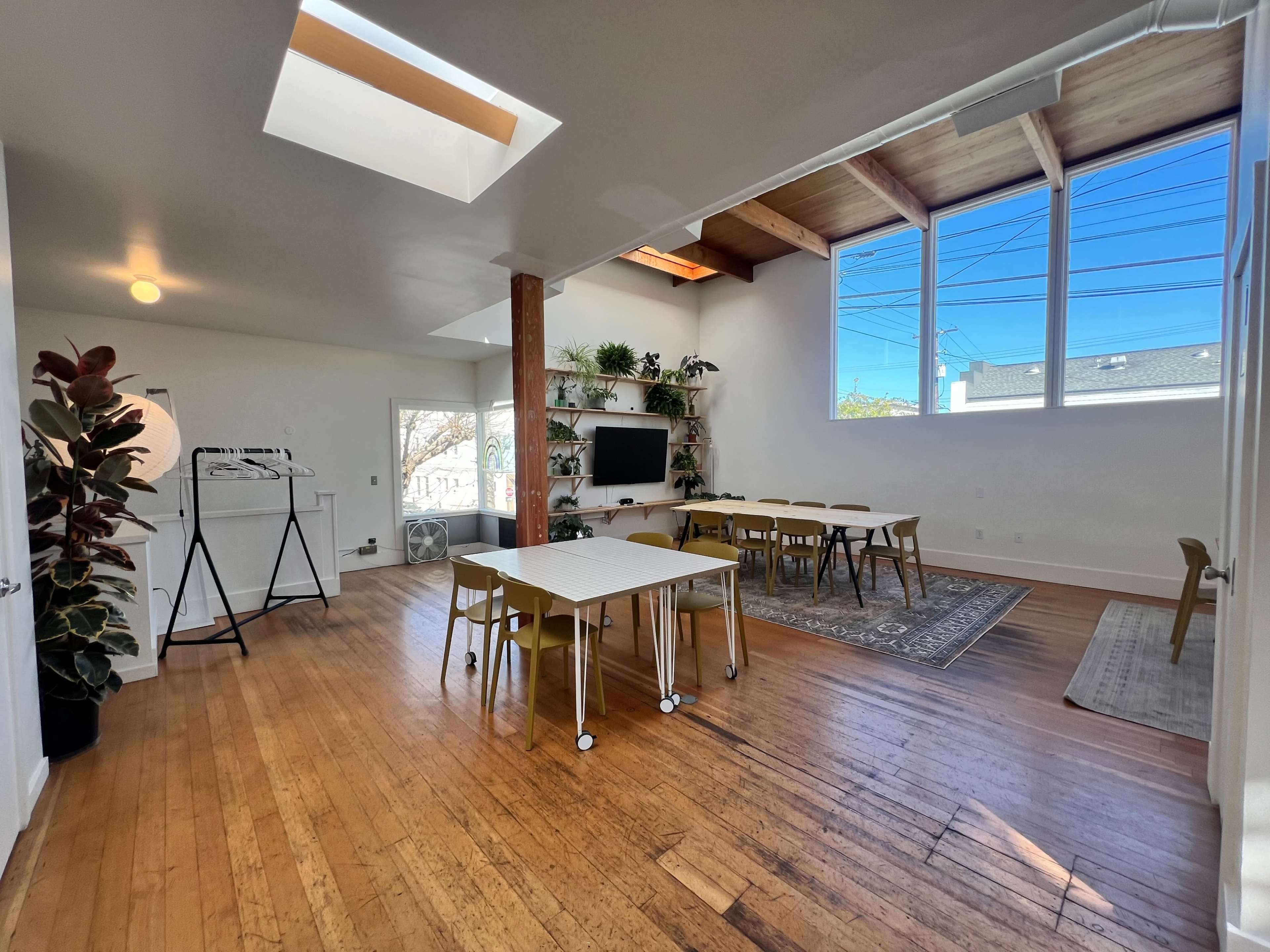 A well-lit, open interior space featuring wooden floors, a large window, tables arranged with chairs, and greenery on shelves.
