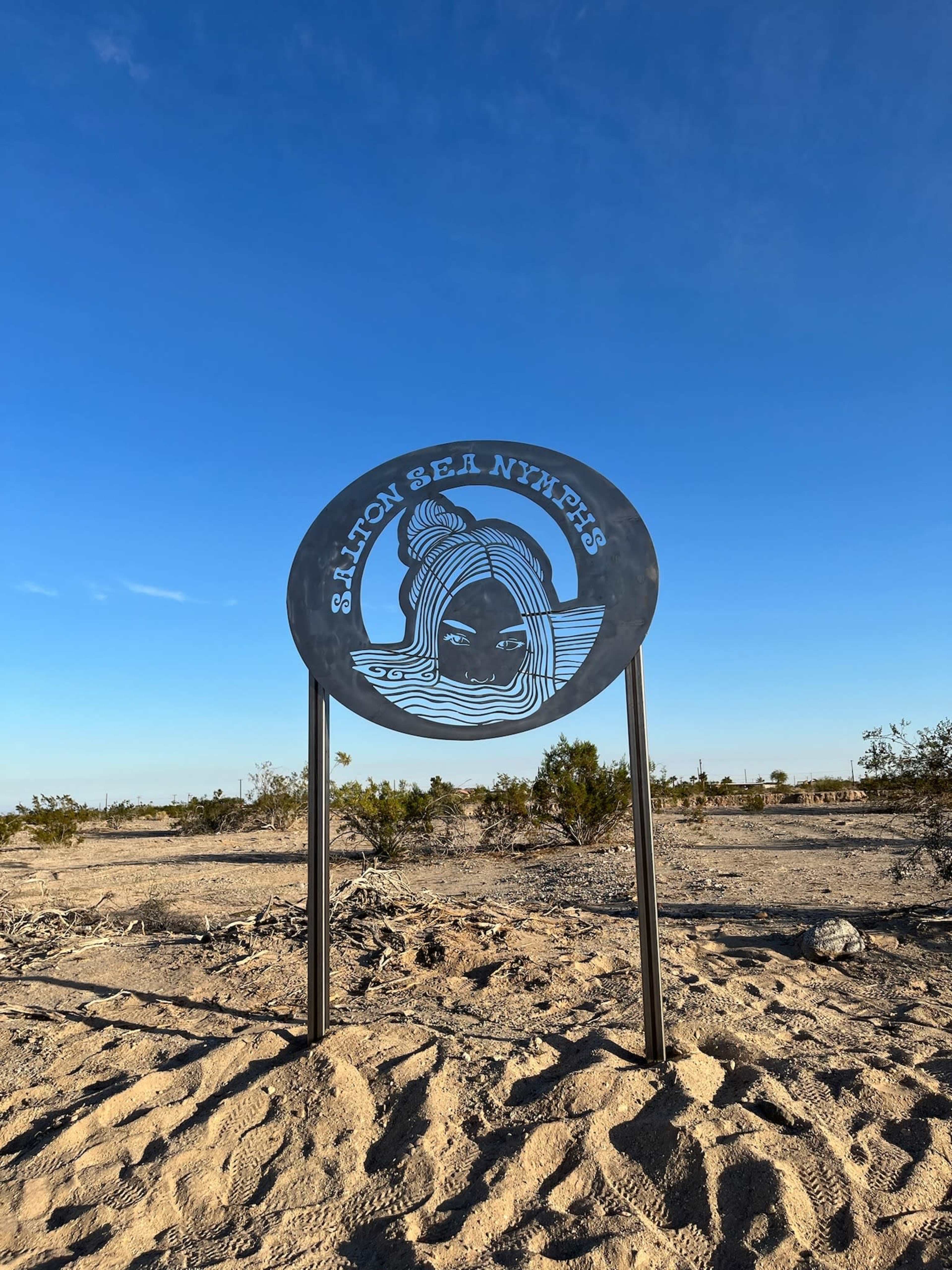 A metal sign displaying "Salton Sea Nymphs" is set against a clear blue sky in a sandy landscape.
