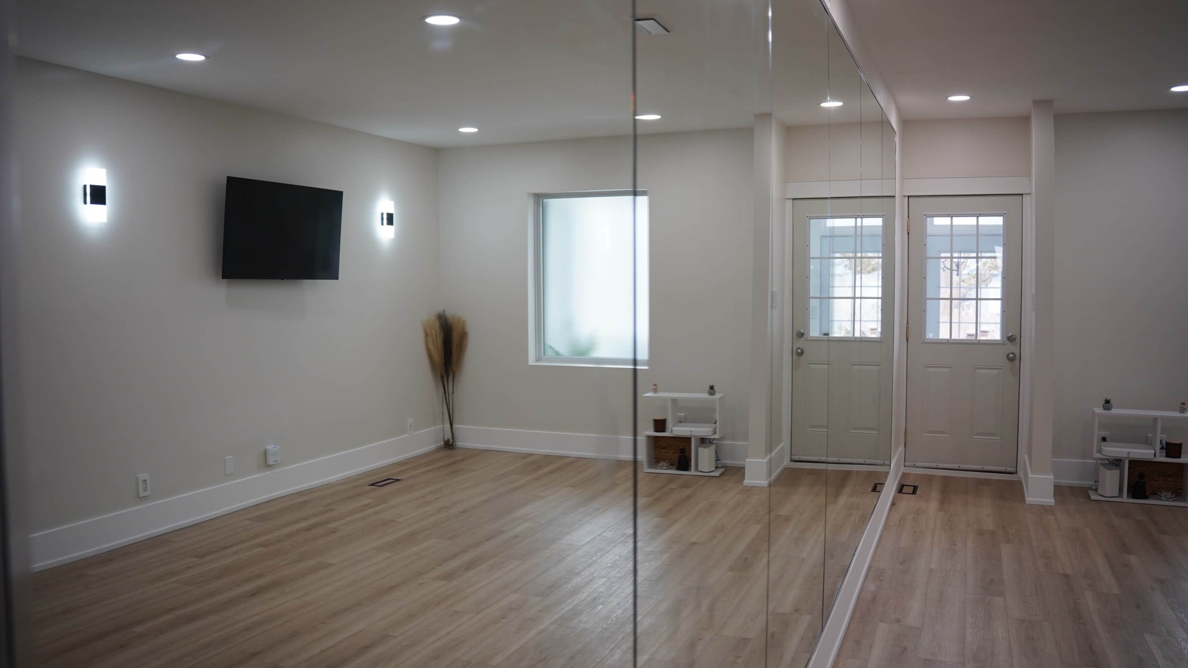 The image shows a spacious, well-lit room with a wall of mirrors, a television mounted on one wall, and a door leading outside.