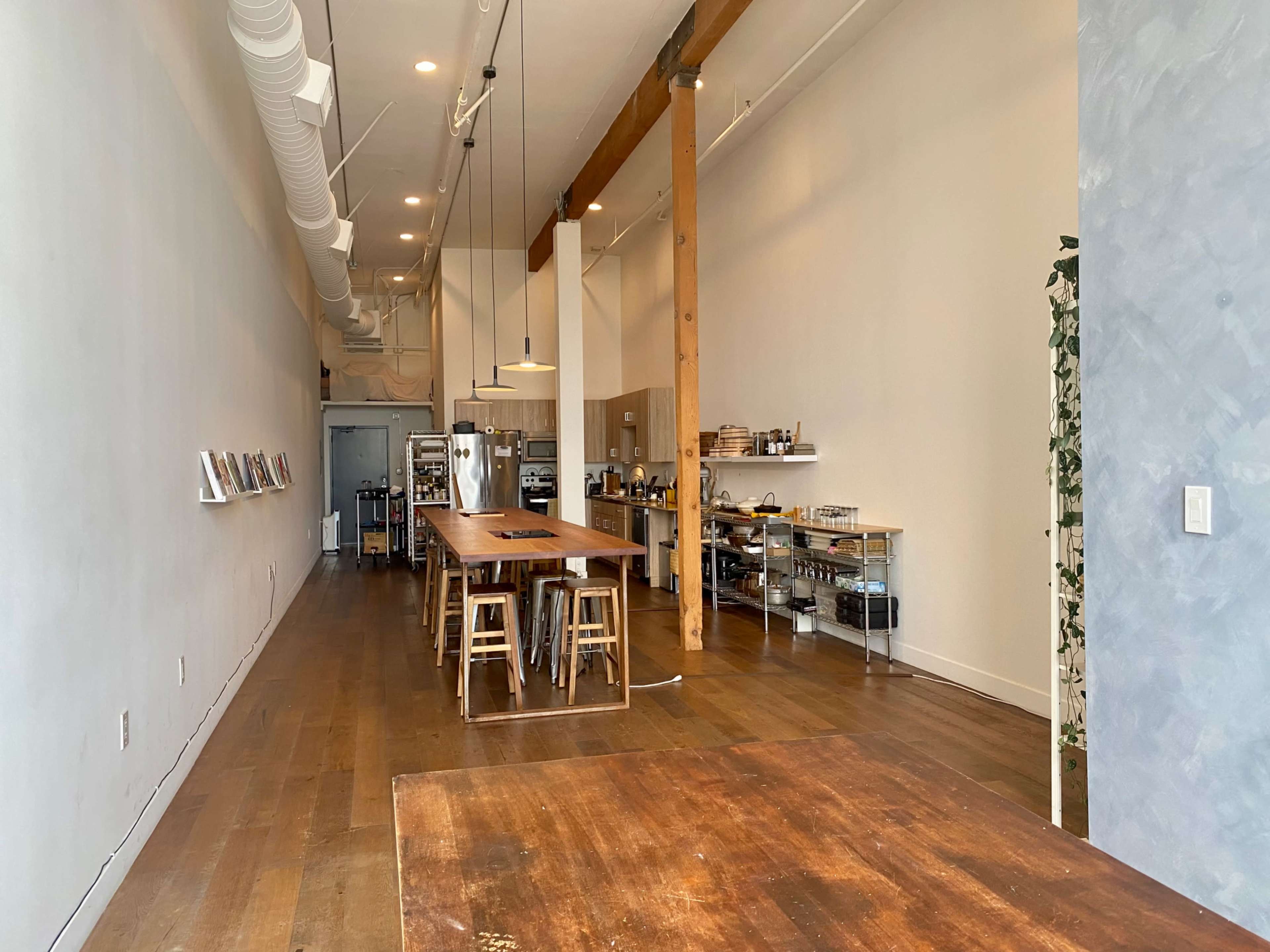 The image shows a spacious, minimalist interior of a café or restaurant with wooden tables, stools, and shelving lined with kitchenware along one side.