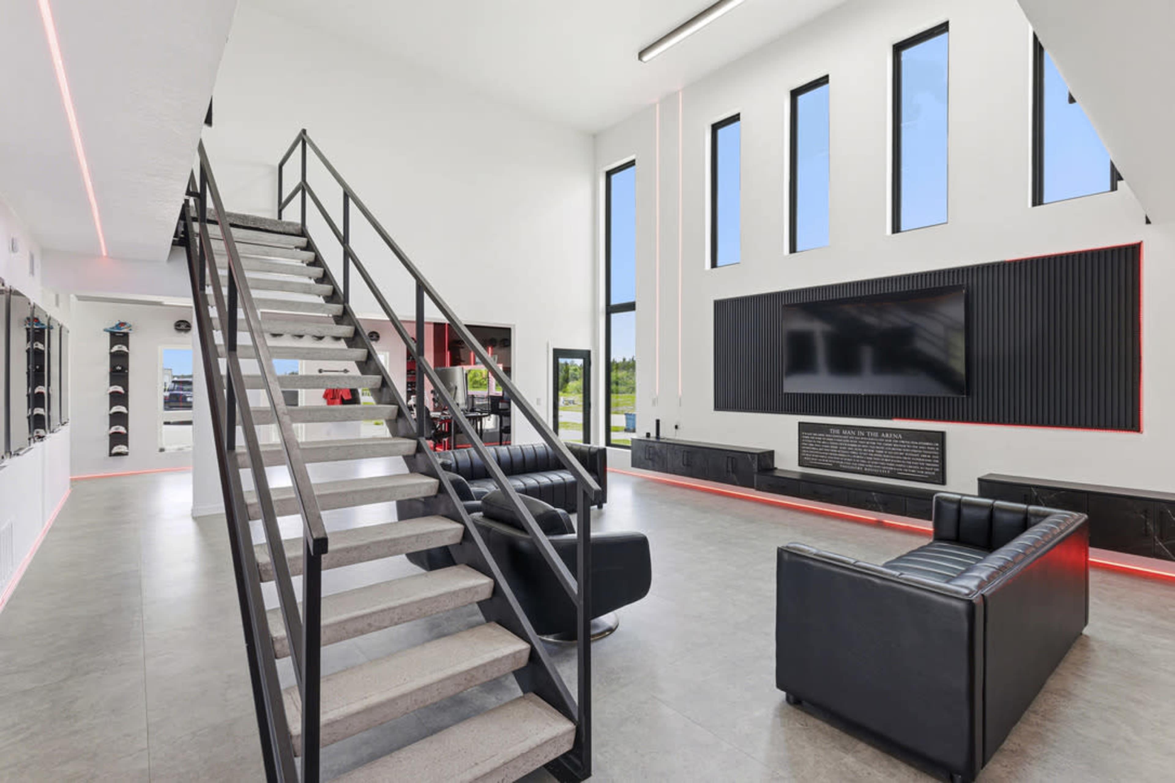 The image shows a modern living space featuring a staircase, black leather sofas, a large television mounted on the wall, and floor-to-ceiling windows.
