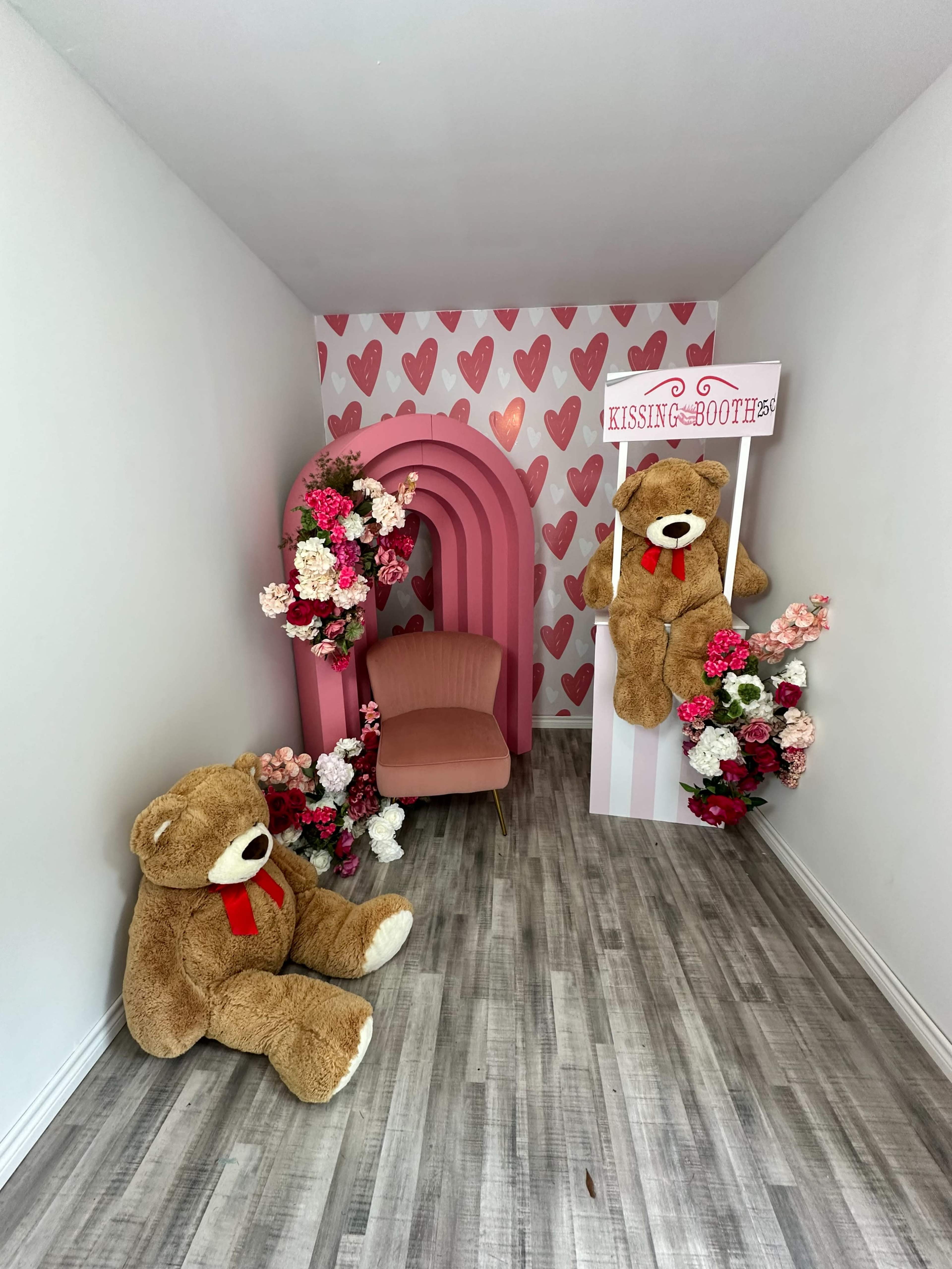 A small room featuring a kissing booth with decorative flowers, two large teddy bears, and a pink chair against a heart-themed backdrop.
