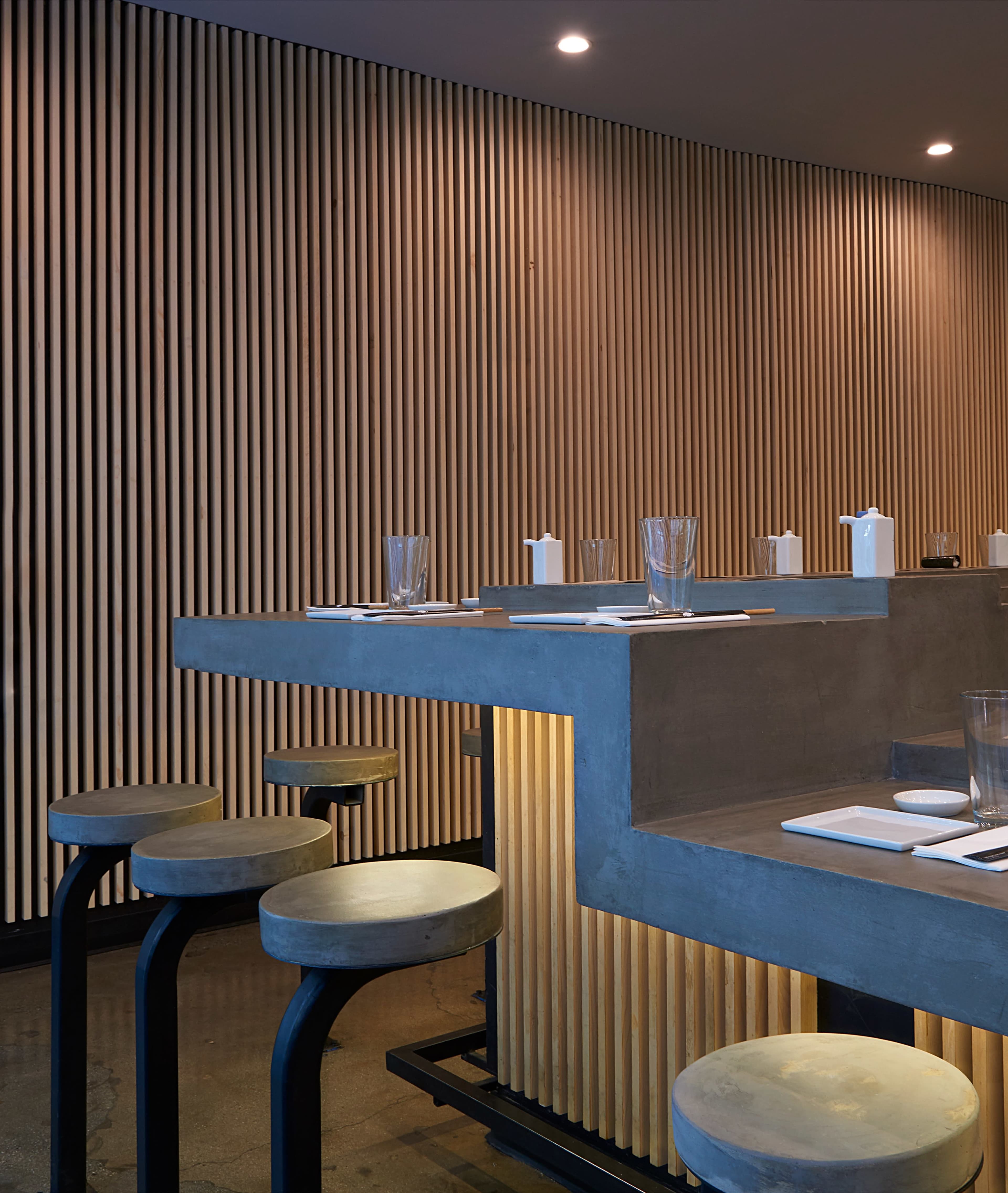 A minimalist bar area with a concrete countertop, wooden paneling on the wall, and circular stools.