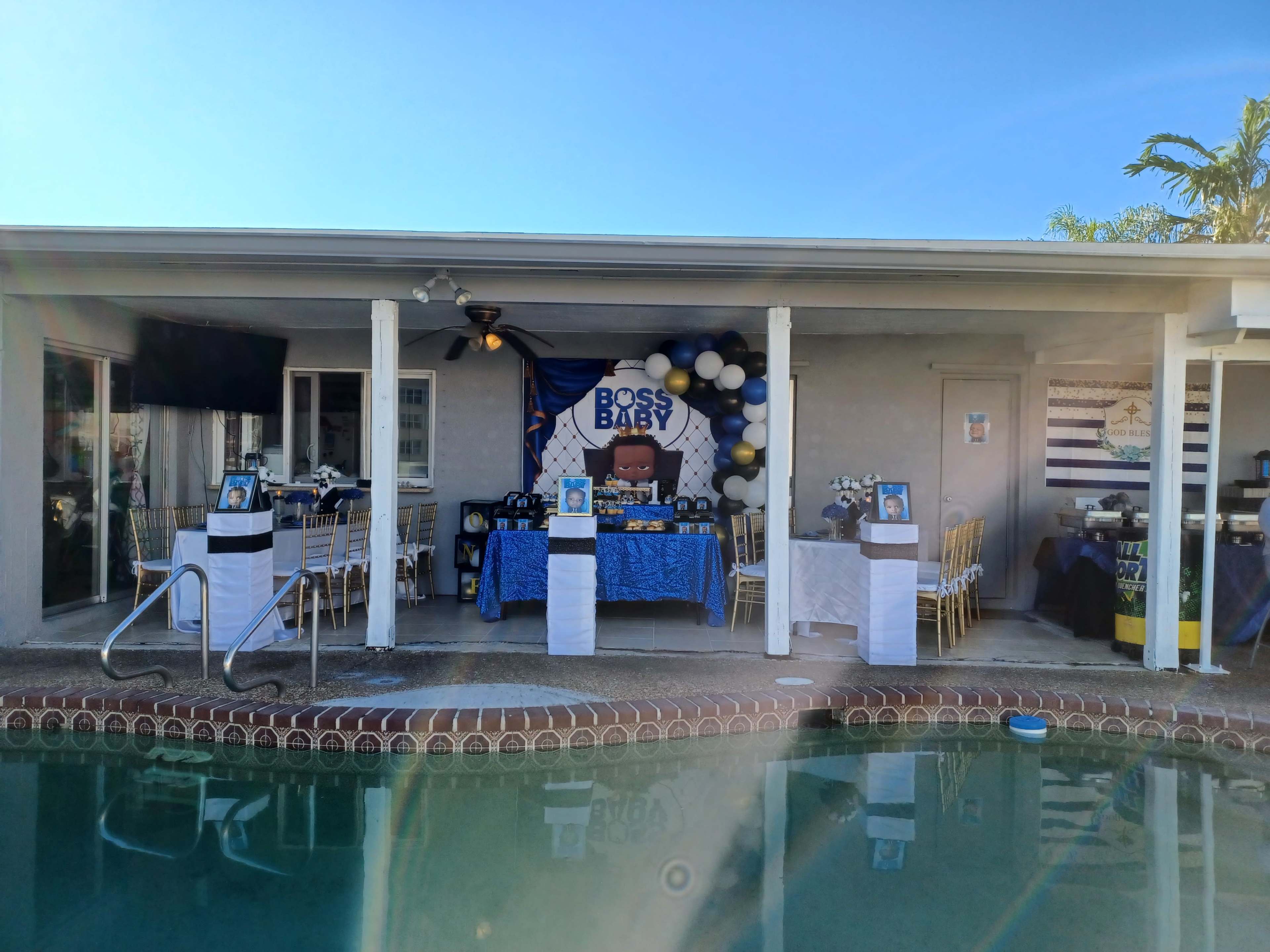 A backyard party setup featuring a "Boss Baby" themed table decorated with balloons, framed photos, and a poolside view.