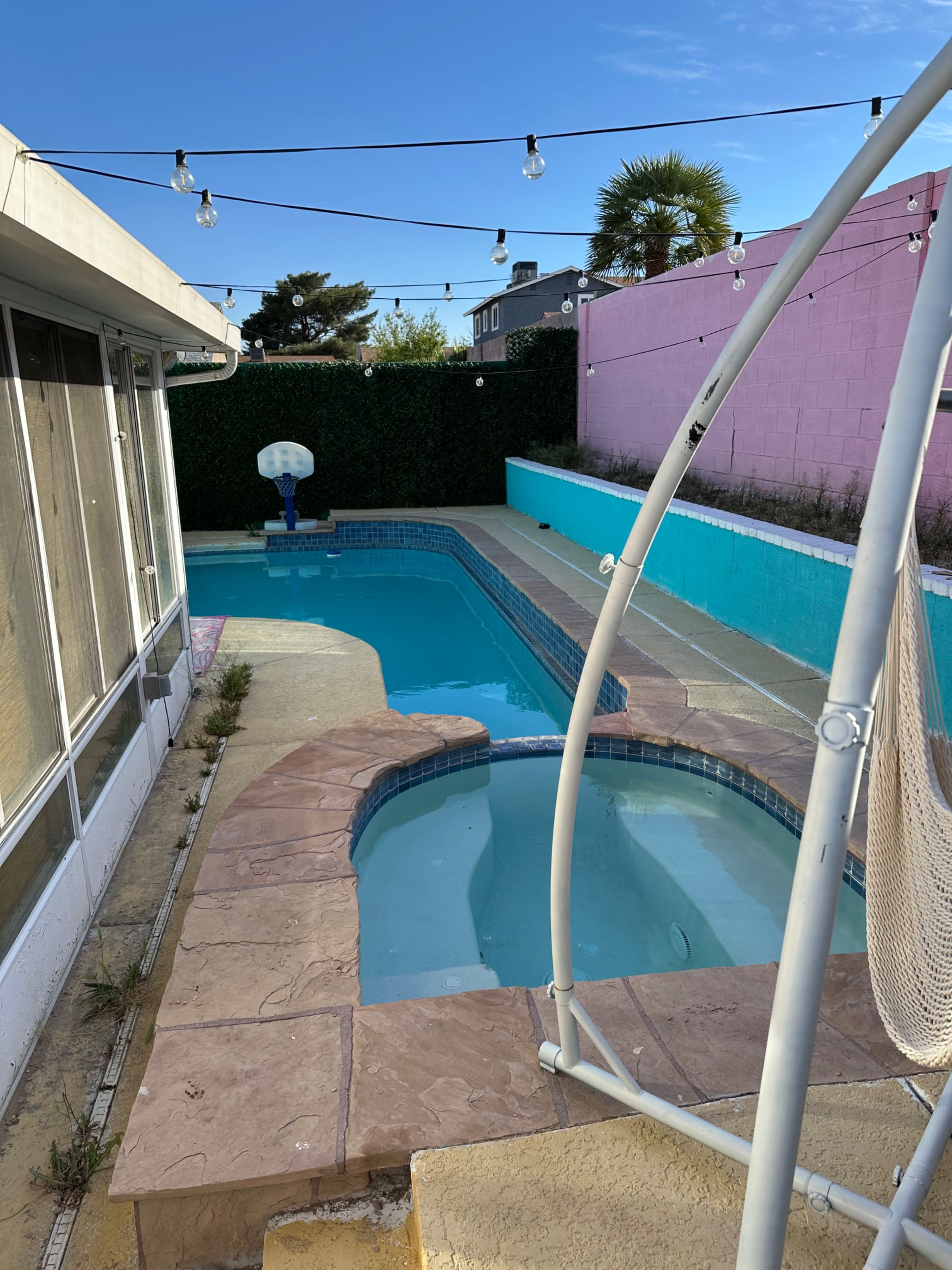 The image shows a backyard pool area featuring a swimming pool, a hot tub, and string lights overhead against a backdrop of green foliage and a pink wall.