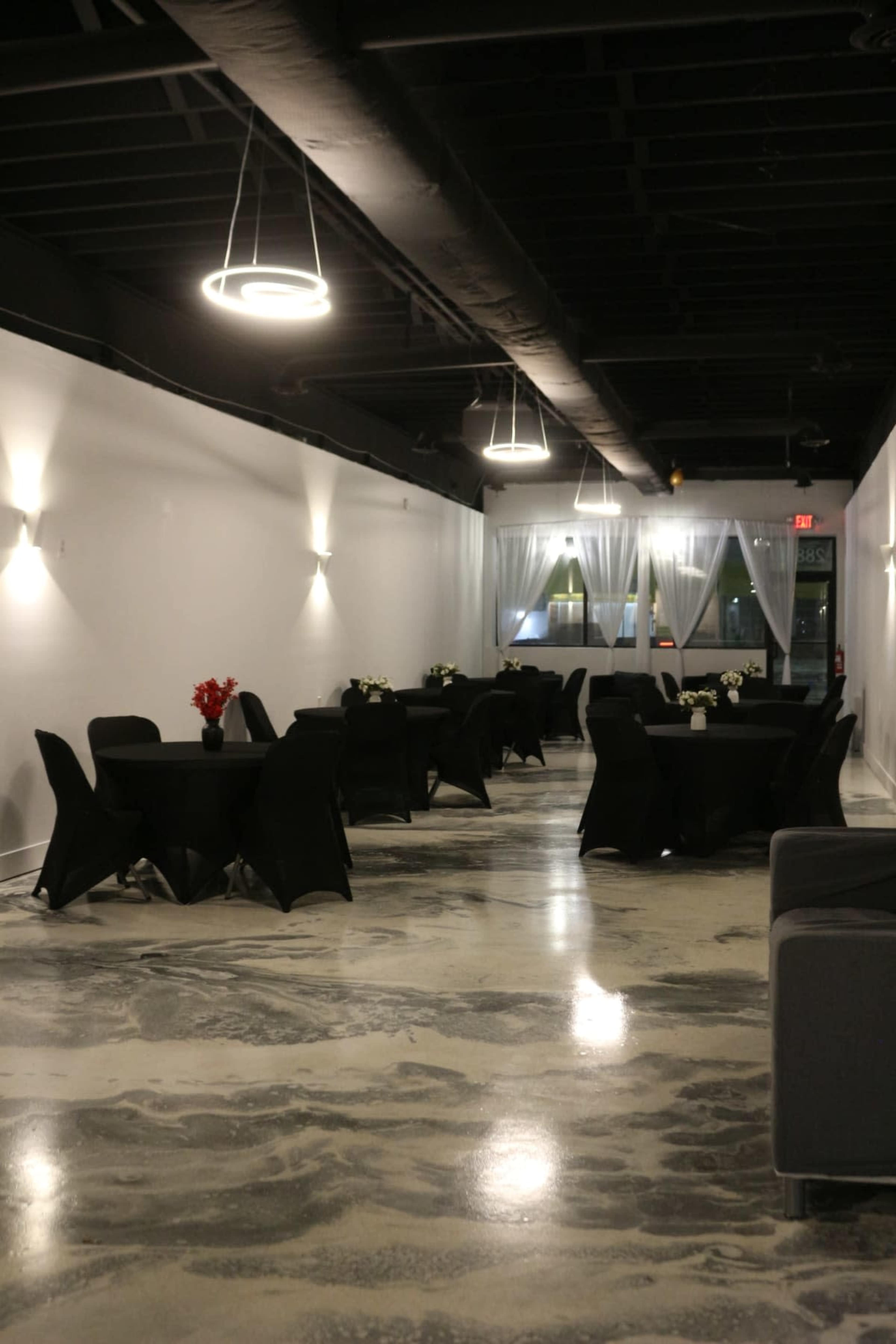 A spacious event room with several round tables covered in black cloth, adorned with floral centerpieces, set against a backdrop of white walls and modern lighting.