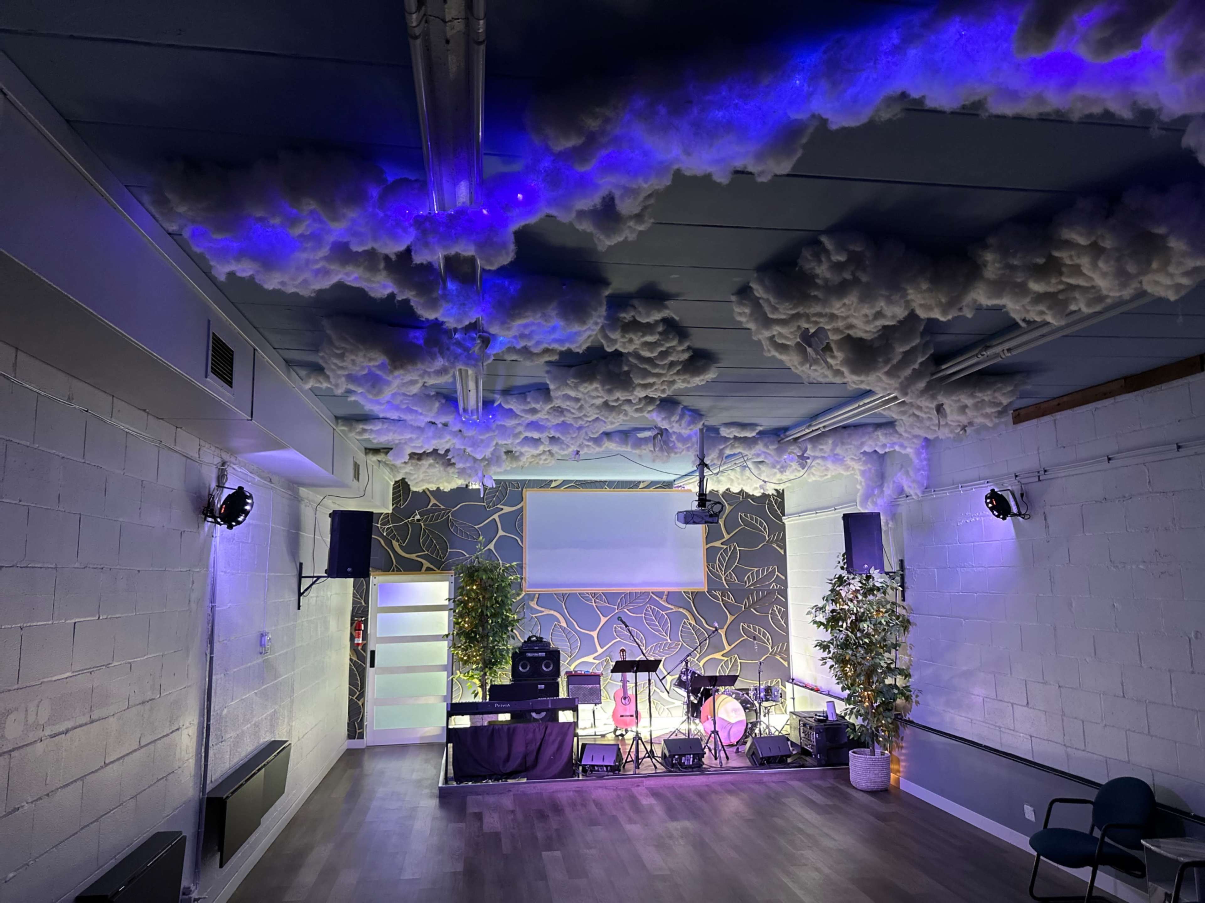The image shows a room designed for events, featuring cloud-like decorations on the ceiling, a stage area with musical instruments, and ambient lighting.