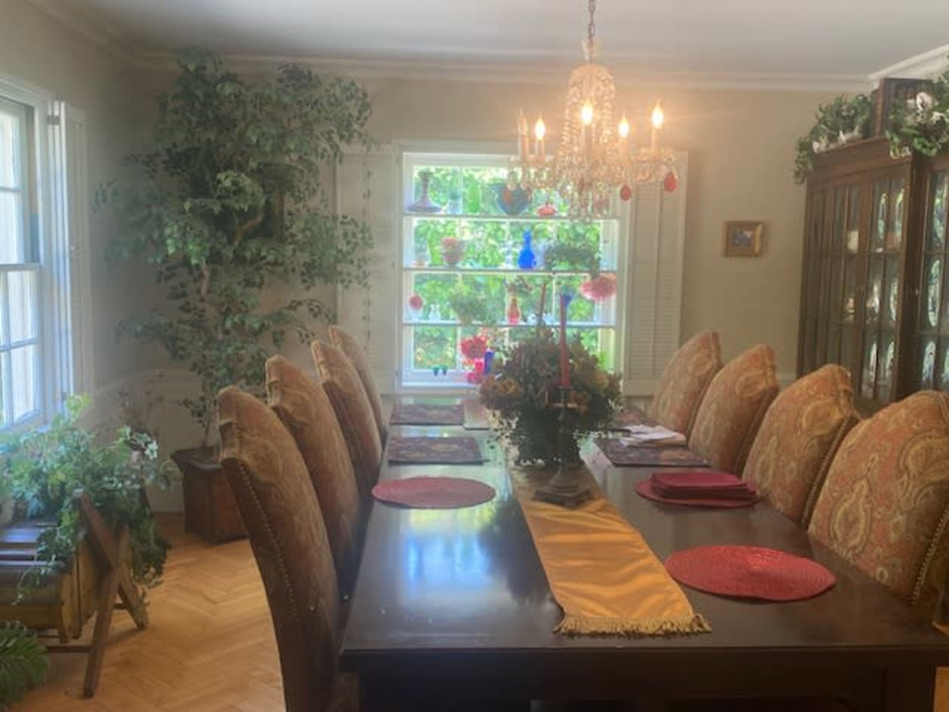 The dining room features a long wooden table set for a meal, surrounded by upholstered chairs, with a chandelier overhead and large windows showcasing greenery outside.