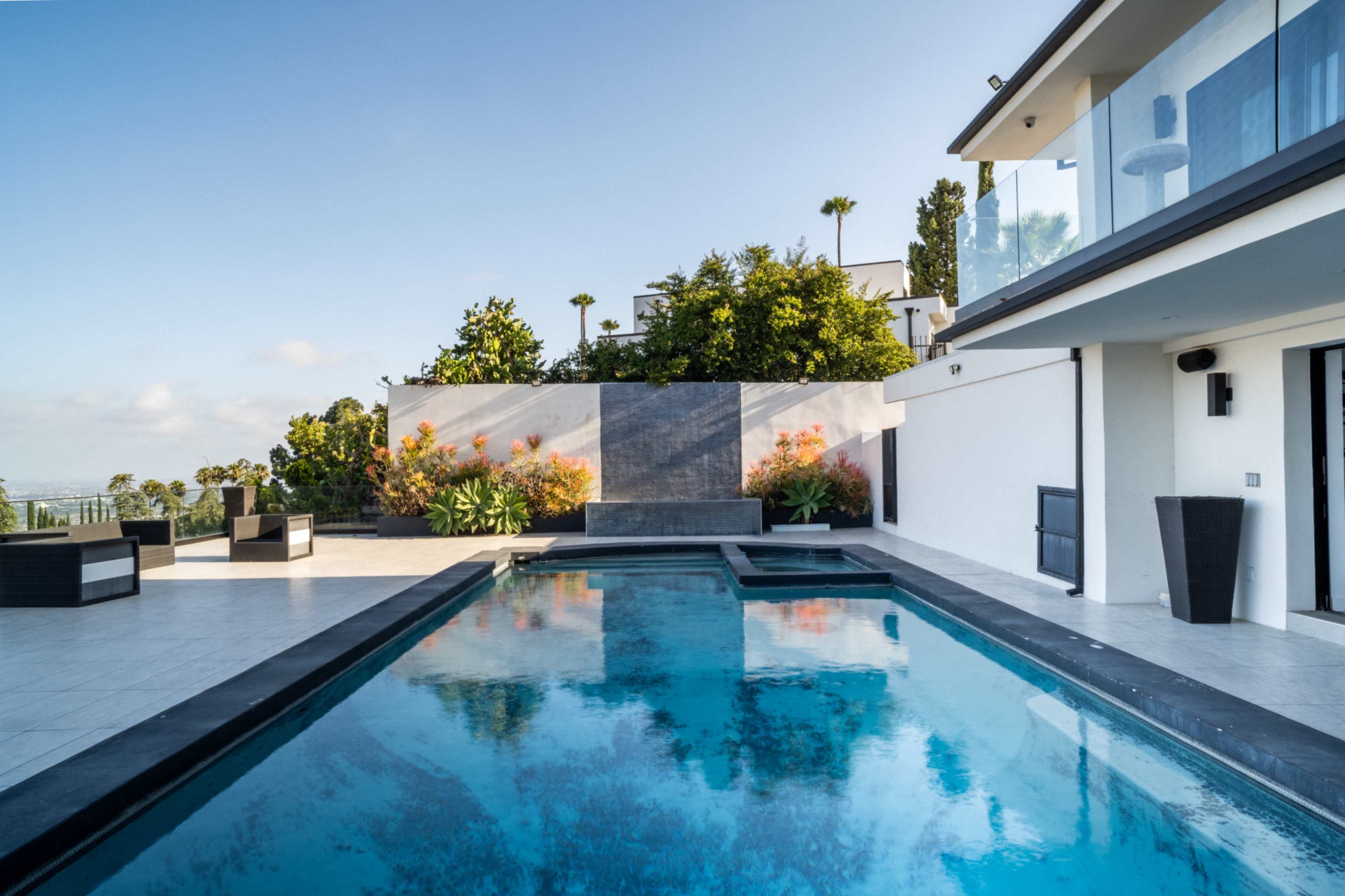 The image shows a modern outdoor area featuring a swimming pool, lounge chairs, and landscaped greenery.